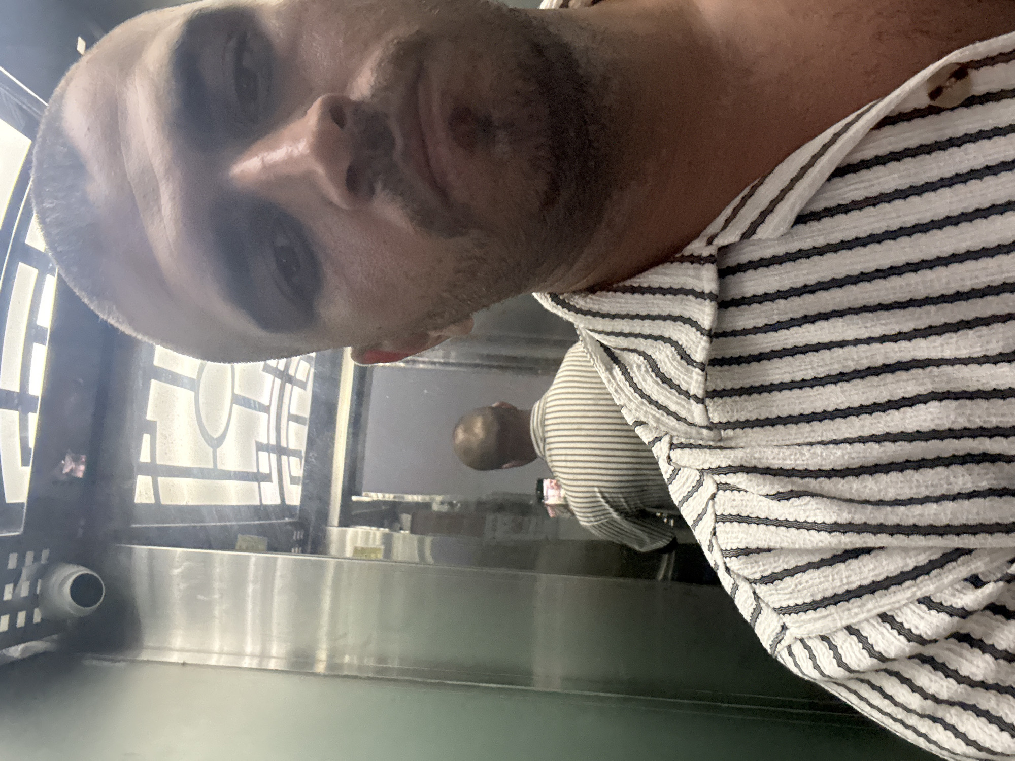 A man is taking a selfie in an elevator. The elevator's interior is silver and gray, with a security camera visible in the corner. A reflection of another person wearing a striped shirt can be seen in the background. The first man is wearing a striped shirt. Đà Nẵng, Vietnam cannot be confirmed.