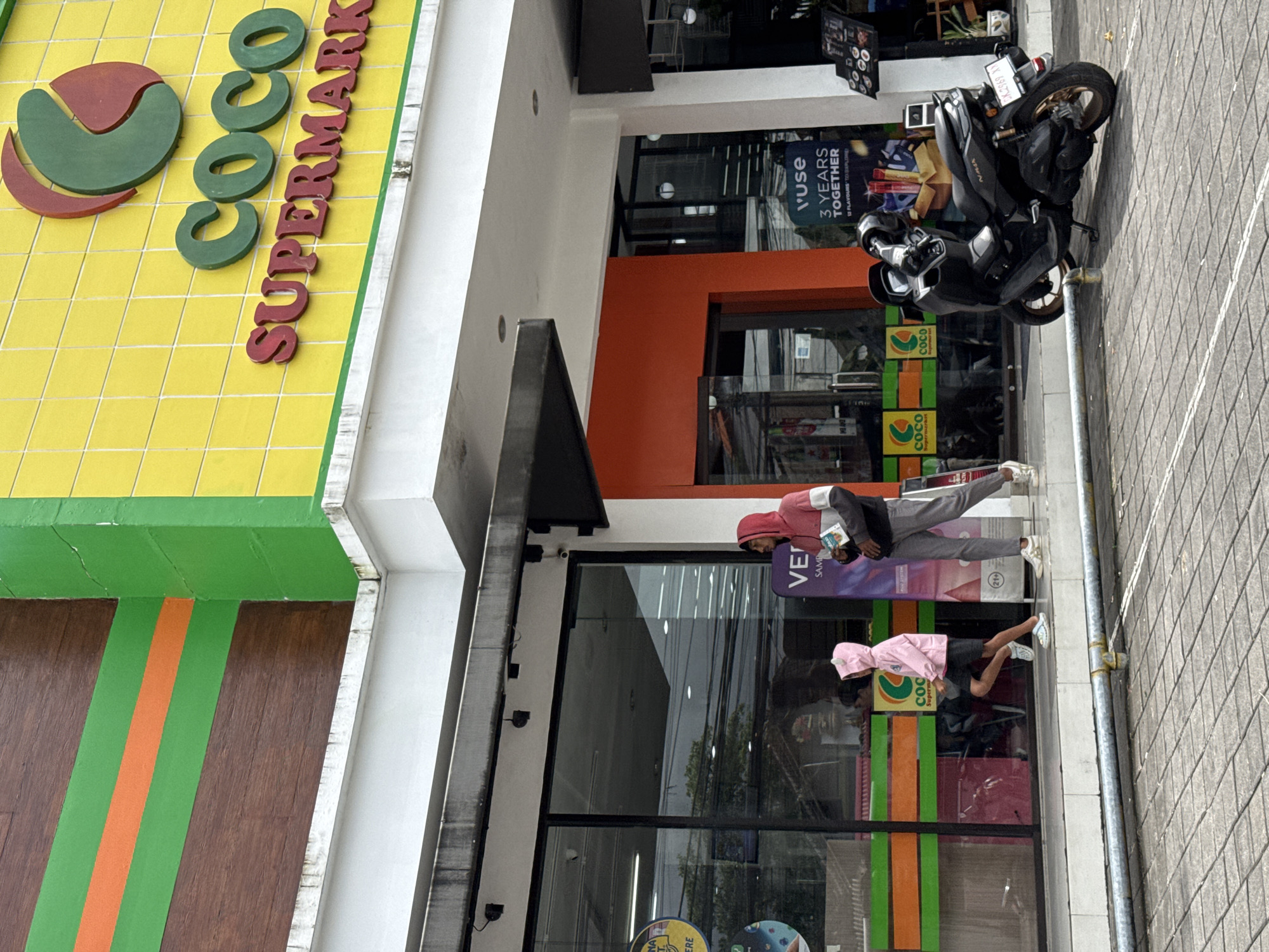 The image depicts the exterior of a Coco Supermarket in Kuta Selatan, Indonesia. The building features a yellow and green facade with the "Coco Supermarket" logo prominently displayed. A motorcycle is parked near the entrance. Two individuals are visible in front of the supermarket. One person is wearing a red hooded top and grey pants, while the other is wearing a pink hooded jacket and black shorts. The environment is urban, with the supermarket fronting a tiled walkway.