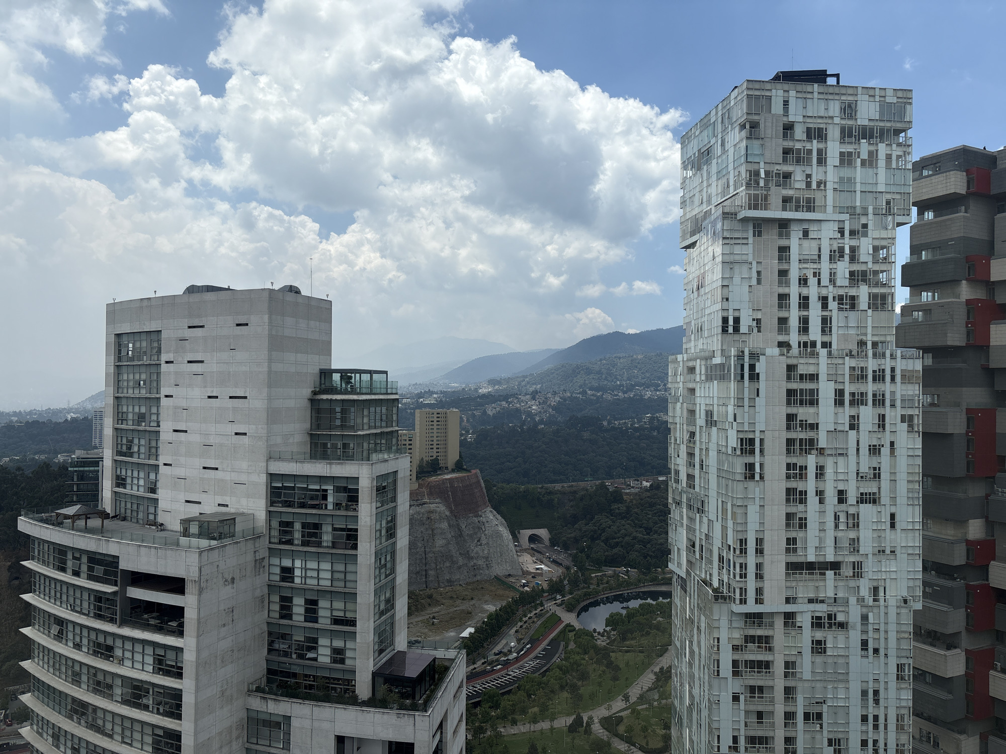 The image presents a daytime view of a cityscape in Mexico City, Mexico. It showcases several modern high-rise buildings with diverse architectural styles. The buildings are predominantly constructed with glass and concrete, featuring geometric designs.
In the background, rolling green hills covered in dense vegetation and residential structures are discernible. The sky is partly cloudy with patches of blue visible amidst the scattered clouds.
Visible between the buildings, a man-made cliff with a tunnel entrance is present, situated near a park with a small body of water and a roadway.