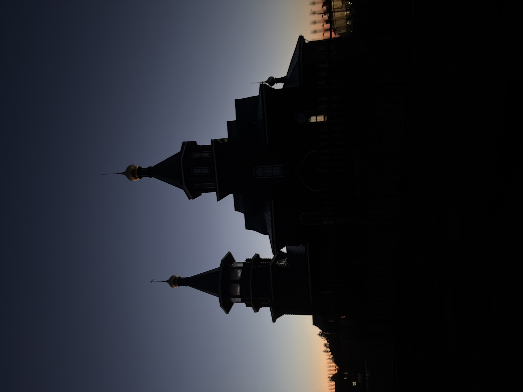The image displays the Church of the Nativity of the Blessed Virgin Mary (Церковь Рождества Пресвятой Богородицы) in Новинки, Nizhny Novgorod Oblast, Russia, presented as a dark silhouette against a twilight sky. The church features two primary structures: a main building with a prominent golden onion dome topped by an Orthodox cross, and an adjoining bell tower to its left, capped with a smaller golden dome and a spire. The architectural details, including multiple tiers and window outlines, are visible despite the deep shadow. A single interior light source illuminates a window on the right side of the main church building. The sky exhibits a gradient, transitioning from a deep indigo at the top to a faint orange and pink hue along the horizon line on the left, indicative of either late dusk or early dawn. The foreground is completely dark, obscuring any ground-level details. Blurred outlines of distant trees or structures are faintly visible on the horizon to both the left and right sides of the church. No people or specific actions are discernible.