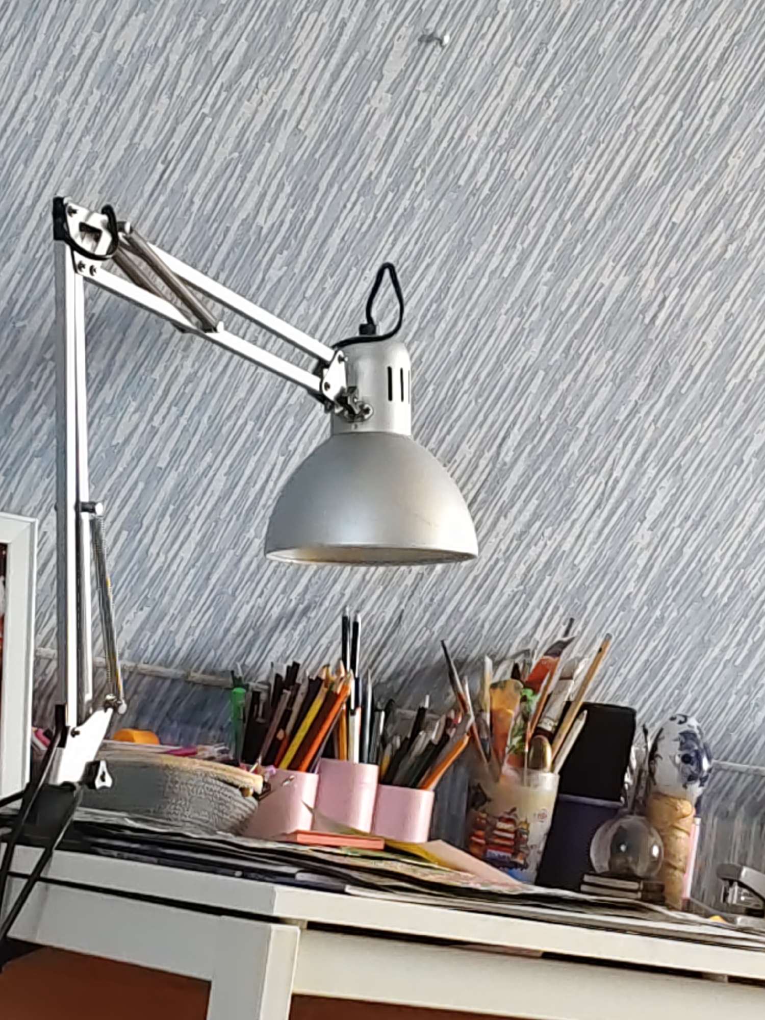 The image shows a close-up of a desk with various art supplies and a desk lamp. The desk lamp, made of metal with a silver finish, is positioned on the left side of the frame, extending over the desk. On the desk, there are several containers holding art supplies, including pencils, pens, and paintbrushes. Some papers and a stapler are also visible. The background is a textured wall with a pattern of diagonal lines. The lighting suggests an indoor setting, possibly a studio or workspace. No people are present in the image. The scene is a still life composition of art-related objects. The specific location in the city of Rostov-on-Don, Russia, cannot be determined from the visual information provided in the image.