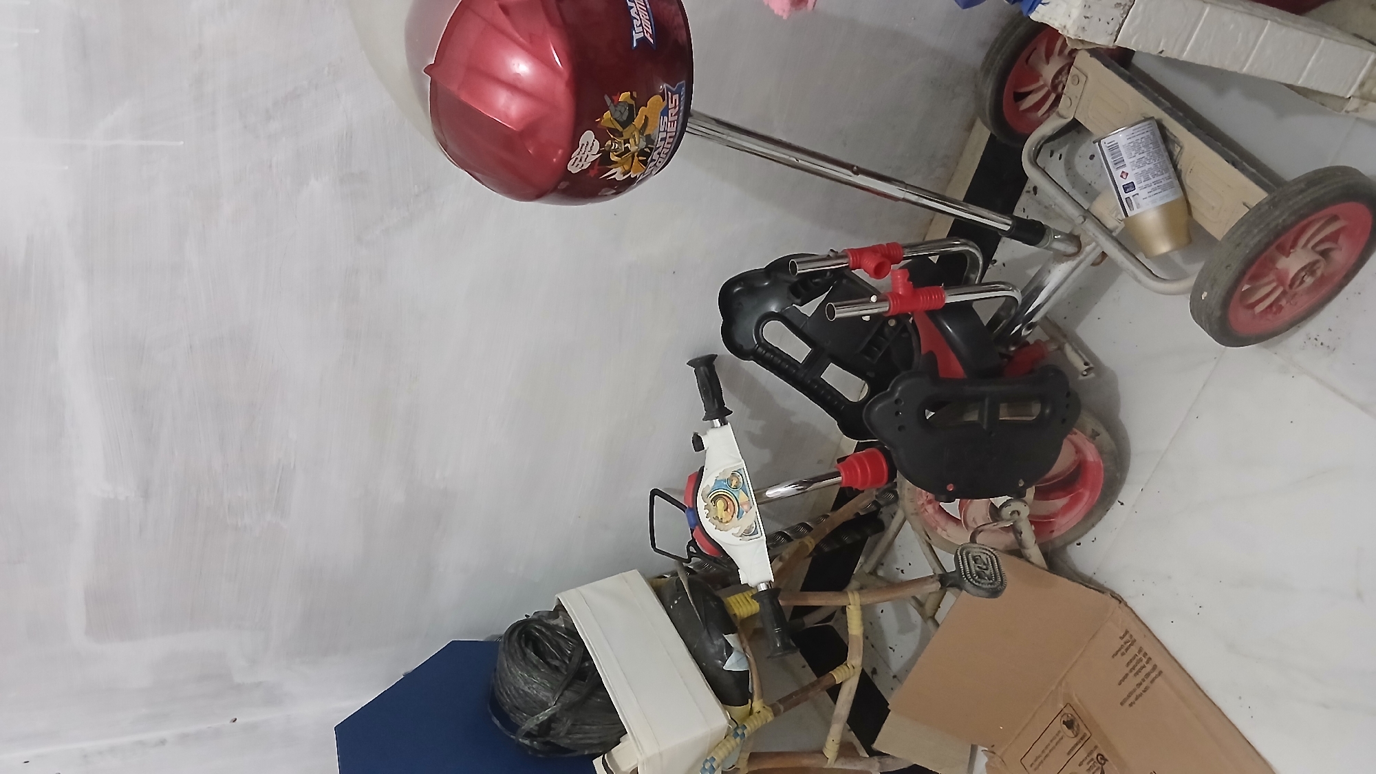 A cluttered indoor scene shows a red bicycle helmet with a Transformers sticker mounted on a silver pole, likely a stand, against a white-washed wall. In front of the helmet, a red and black children's tricycle is partially visible, with some of its parts including wheels, handlebars, and footrests. To the left of the tricycle, a cane chair with a white cloth basket filled with black plastic rolls is placed. A cardboard box is visible on the floor in the lower left corner of the image. The floor is tiled in a light color, and the overall environment suggests a storage or unused area. The context of Klambir Lima Kebun, Indonesia, is not visually evident in the image.