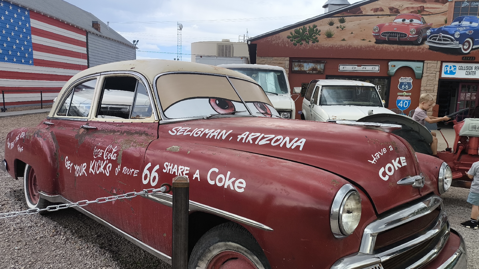 An aged, red Chevrolet sedan, decorated with cartoonish eyes on the windshield, is parked on a gravel lot in Seligman, Arizona, USA. White lettering on the car's hood and sides reads "SELIGMAN, ARIZONA," "GET YOUR KICKS ON ROUTE 66," "SHARE A COKE," and "Have a COKE." The car is chained off. In the background, a building with a mural of classic cars and a Route 66 sign is visible. A young boy is sitting on the hood of another vehicle, holding onto a steering wheel.
