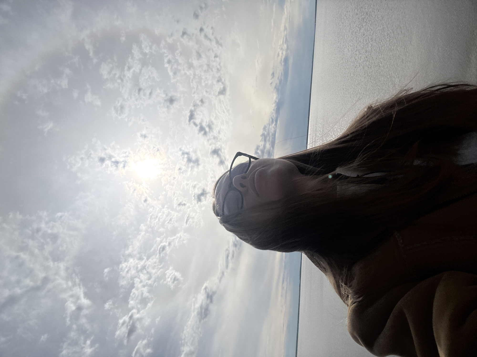 A person with long brown hair and glasses is looking up towards a bright sky with scattered clouds. The sun is visible through the clouds, creating a halo effect. The person is wearing a tan-colored garment. In the background, there's a body of water that could be a lake or sea, appearing flat and reflective. The location is Timiryazevskii S/S, Ivanovo, Russia.