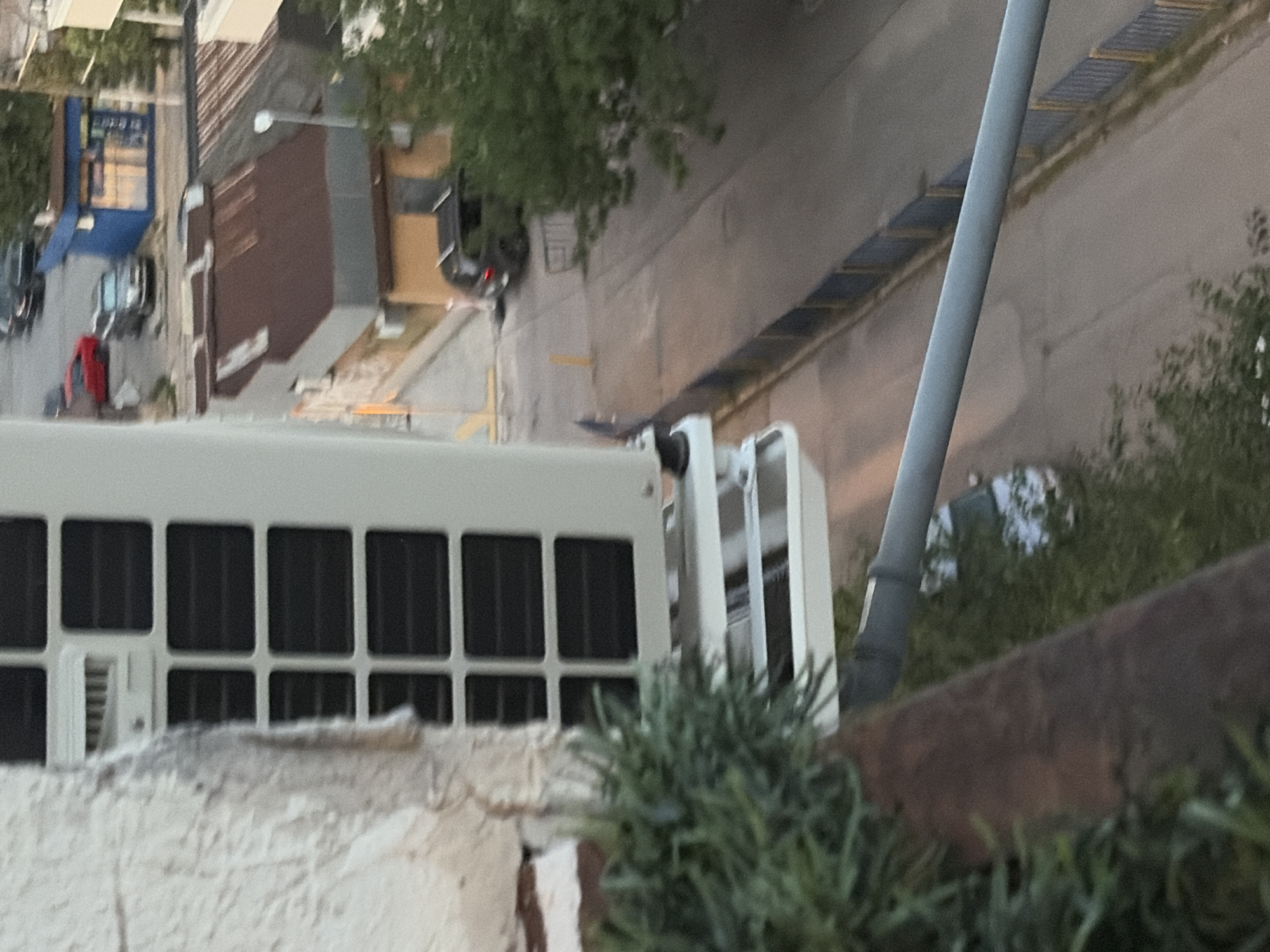 An outdoor, eye-level, vertical shot captures an air conditioning unit mounted on a textured, off-white wall. The unit itself is white and features a grid of black rectangular vents. Below the main unit, a set of horizontal white slats is visible. To the left, a dense green shrub obscures part of the image. In the background, a street scene unfolds. A dark-colored SUV is parked on the right side of the street. Further down the street, a small, blue building with windows is situated. Several other vehicles, including a red car, are also present on the street. A portion of a rusty, sloped roof of a building is visible in the middle ground. A grey pipe runs horizontally across the lower portion of the frame, partially obscuring the view of the street below. The lighting suggests it may be late afternoon or early evening.