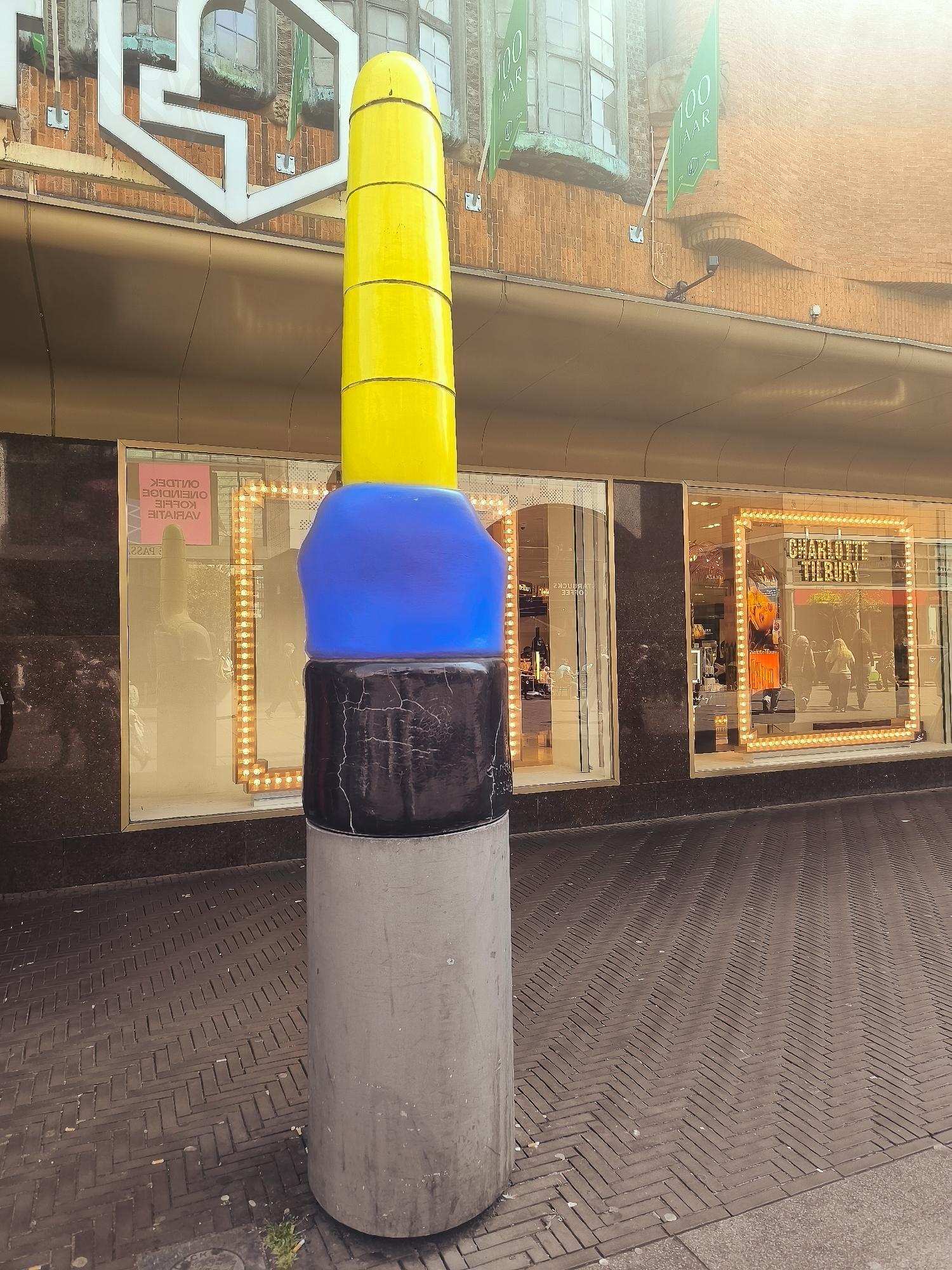A tall, cylindrical sculpture is positioned on a brick-patterned sidewalk in front of a store. The sculpture is segmented into three parts: a gray, concrete-like base, a black section with visible cracks, and a bright blue section. Atop the blue section is a tall, tapered yellow section with a rounded top. Behind the sculpture, a store window displays mannequins and is illuminated by numerous small lights arranged in a rectangular frame. The store facade has a reflective, gold-colored awning. Above the awning, the building is brick, with visible architectural details and windows. Green banners with the text "100 JAAR" (100 Years) are displayed on the upper part of the building. The overall scene suggests an outdoor urban environment in The Hague, Netherlands, likely in a shopping district.