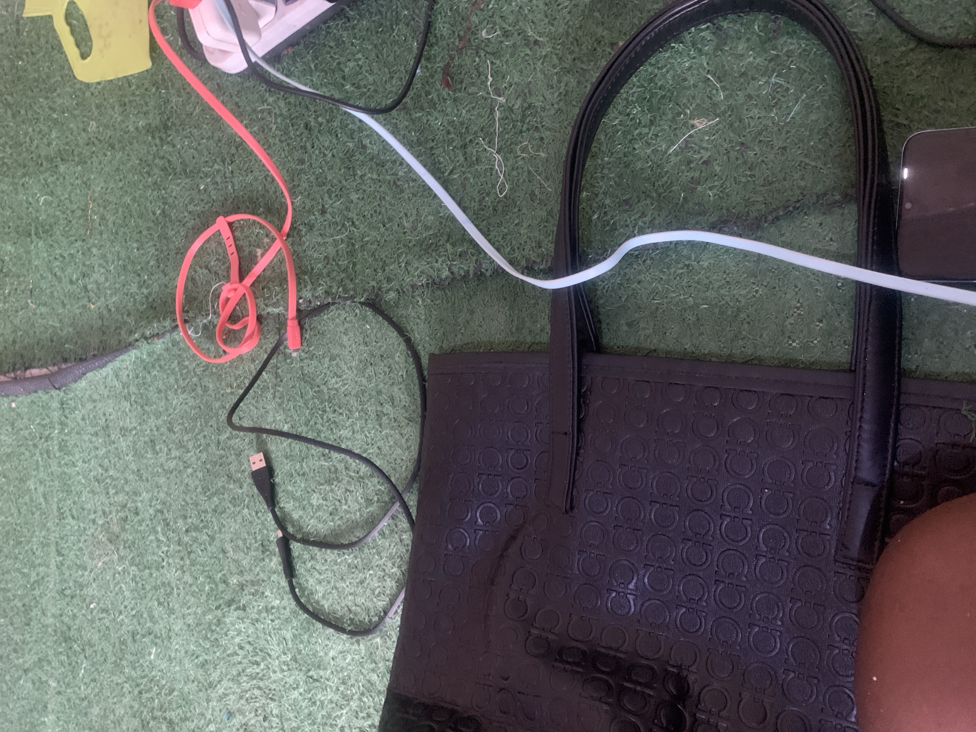 The image depicts a close-up of a black handbag with an embossed pattern, lying on a green, carpet-like surface. Several charging cables are scattered around the handbag, including a black USB cable, a coiled pink cable connected to a power strip, and a white cable. A portion of a person's leg, with darker skin tone, is visible in the lower left corner of the frame. A smartphone is partially visible in the lower right. The scene appears to be an indoor or outdoor setting with artificial grass or a similar textured ground cover. The location is identified as Jauro Yinu, Nigeria.