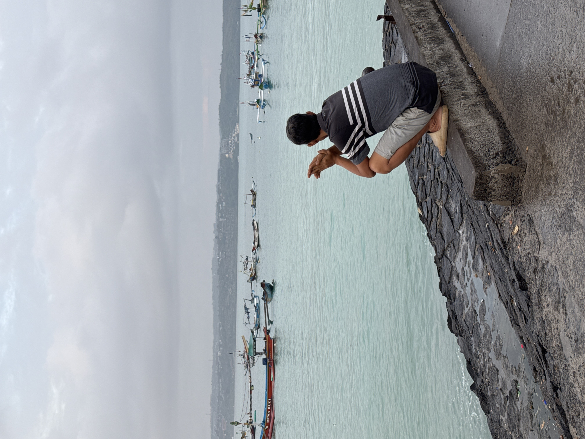 A man wearing a striped shirt and light-colored shorts crouches on a concrete barrier overlooking the ocean. The man is facing away from the camera, his hands clasped in front of him. The water is a turquoise color, and several traditional Balinese fishing boats are visible in the distance. The sky is overcast, and the overall atmosphere of the image is serene. The scene is likely set in Kuta, Indonesia, a popular tourist destination known for its beaches and vibrant fishing culture.