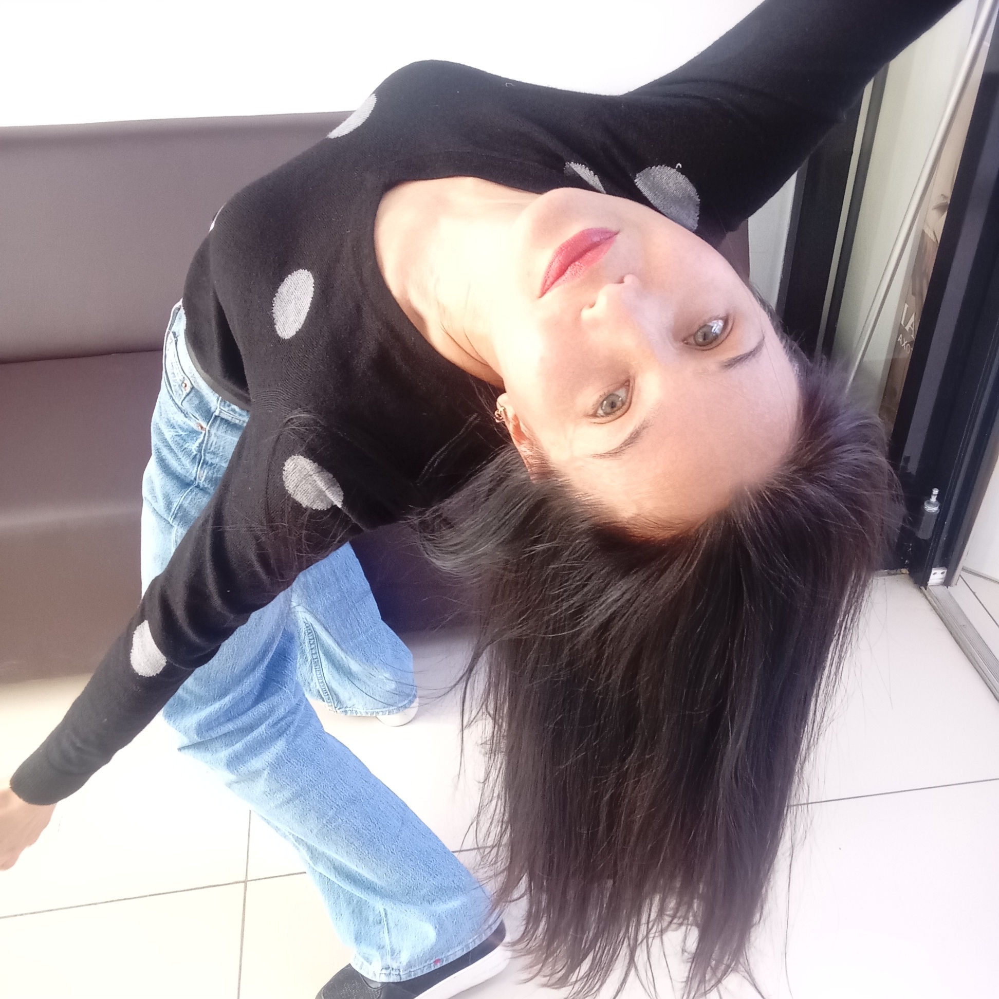 An individual is captured in a dynamic, full-body portrait, leaning significantly backward with their body angled and head tilted upwards, looking directly into the camera. The subject is a woman with long, dark, straight hair that cascades downwards due to her posture. Her eyes are light-colored, and she wears red lipstick.
She is dressed in a black long-sleeved top adorned with large, light grey or white polka dots, and light blue denim jeans. Her arms are extended outwards, contributing to the posed nature of the photograph. Black sneakers with white soles are visible on her feet.
The setting appears to be an indoor public space, possibly a waiting area, lobby, or a retail environment in Izhevsk, Russia. A dark brown or grey sofa or bench is visible behind her to the left. The floor is covered with large, light-colored tiles. To the right, there's a glimpse of a display or glass panel, possibly a shop window or door. Faint, partial text is visible on this panel, including fragments that look like "LAX" and "AXO". The lighting is bright and even, suggesting an indoor light source. The overall impression is a casual but artfully composed shot.