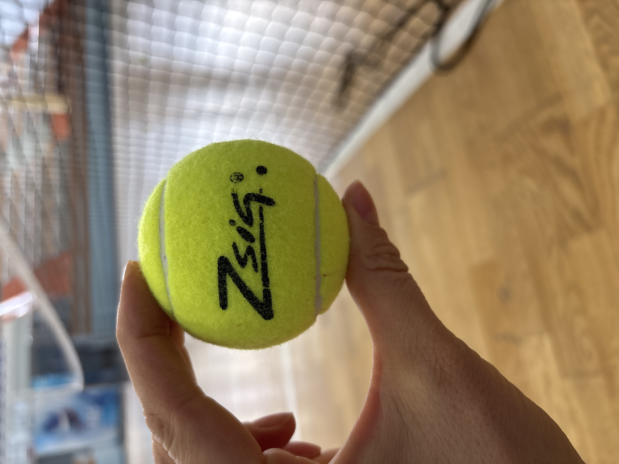 A human hand, primarily showing a thumb and index finger, grips a fluorescent yellow tennis ball with a visible black "ZSIG." logo. The skin tone of the hand is light. The background is out of focus, revealing a white mesh tennis net stretched across the mid-ground. Below the hand and ball, a light-colored wooden court floor is visible. Distant blurry elements behind the net suggest an indoor facility with various light-colored structures and a blue object. The scene takes place within a sports hall in Chaikovsky, Russia.