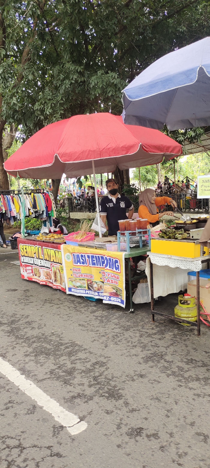 The image shows a street food stall in Kecamatan Sidoarjo, Indonesia. The stall is located under a red and a blue umbrella, with a few people around. The stall offers a variety of food items, including fried chicken, fried fish, and "nasi tempong". One man is standing near the stall, wearing a blue shirt with the number "VIII" on it, while a woman in a yellow shirt with a green headscarf is preparing food behind a counter. In front of the stall, there are a few tables covered with a white table cloth, and some metal chairs. On the counter there are several containers of food and drinks, including what appear to be some skewers of chicken, a yellow container filled with food, and several glass jars filled with liquid. The street is paved with asphalt, with a white line painted on it. Behind the stall is a rack filled with clothes, a metal fence, and a row of green trees. The surrounding area is bustling with activity, with people walking by, and other food stalls nearby.