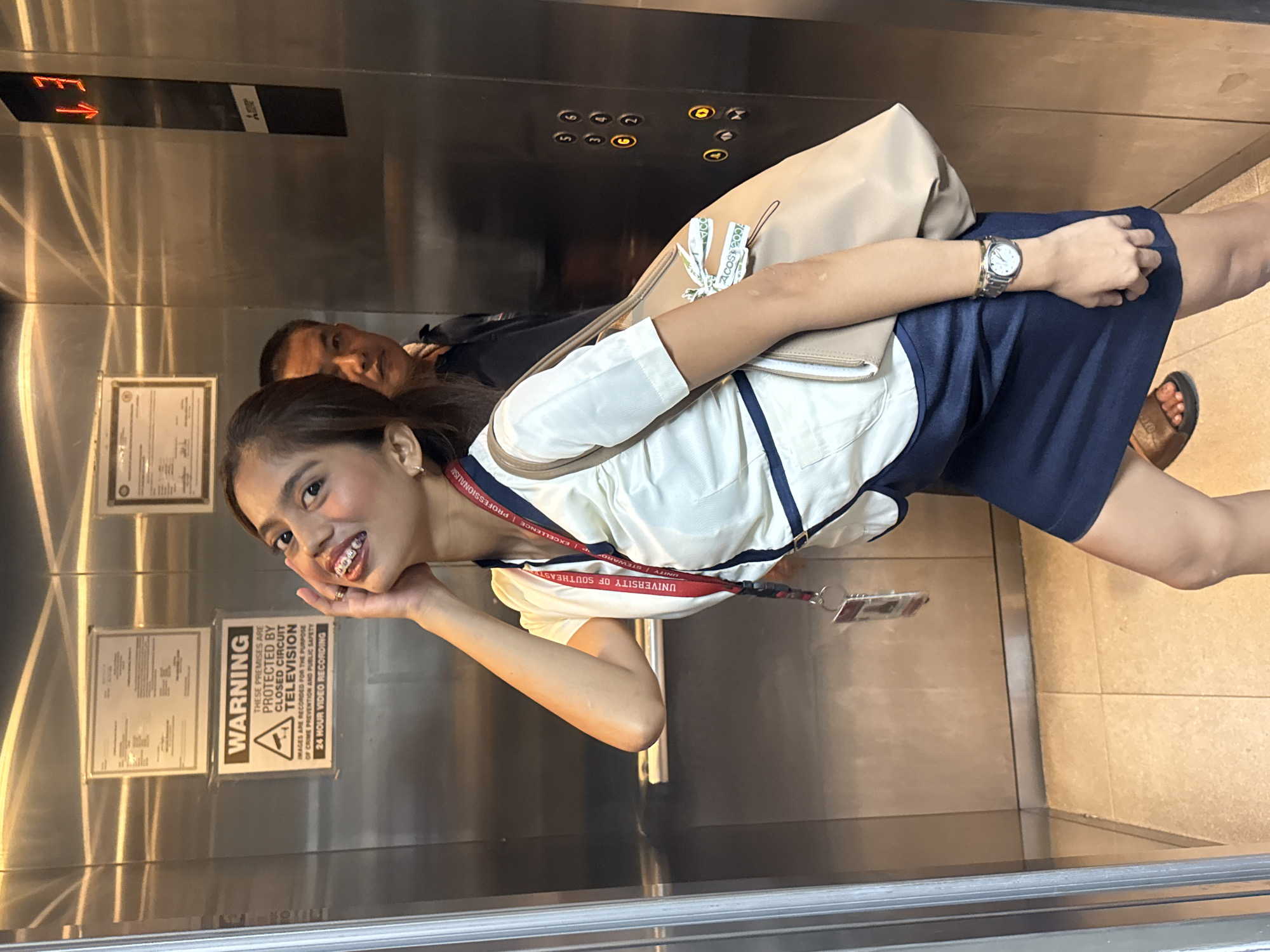 A woman with braces and dark hair, wearing a white collared shirt, a dark blue skirt, and a lanyard, poses in an elevator. She is holding her right hand to her face and looking at the camera with a smile. A beige bag is slung over her left shoulder, with a white ribbon attached. Behind her, a man with short dark hair and a dark shirt stands facing the elevator doors. The elevator walls are made of brushed metal, and there are signs on the left wall, one of which reads "WARNING THESE PREMISES ARE PROTECTED BY CLOSED CIRCUIT TELEVISION". The elevator floor is tiled in a light beige color. The elevator panel shows buttons labeled with numbers and symbols, and an illuminated display shows "1 3" with red downward-pointing arrows. The scene appears to be inside an elevator in a building located in Davao City, Philippines, based on the implied context of the "University of Southeast Asia" branding on the lanyard, which aligns with institutions in that region.