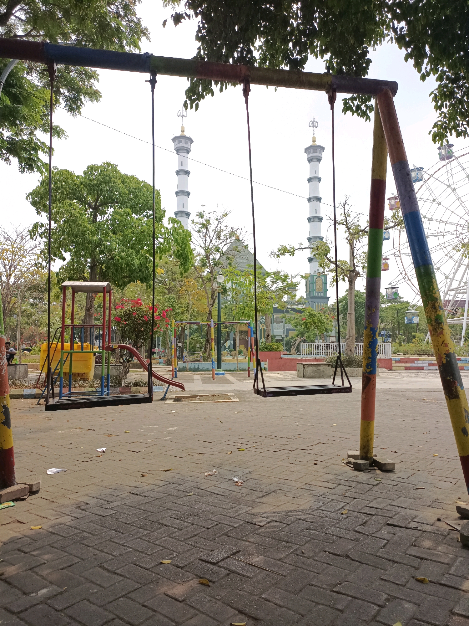 This is a medium-shot, eye-level, outdoor photograph taken in what appears to be a park or playground area in Lamongan, Indonesia. The foreground is dominated by a colorful, weathered swing set with two black swings hanging from it. The swing set's support poles are painted with sections of red, yellow, blue, and green. The ground is paved with interlocking gray bricks.
In the middle ground, there is a small children's play structure with a yellow slide and a red climbing frame. Behind the playground equipment, trees with green and some yellowing leaves are visible, indicating a possible autumn season. Further in the background, two tall, slender minarets of a mosque rise into the sky. The mosque itself has a green dome and architectural details. To the right of the minarets, a large Ferris wheel is partially visible, with several colorful gondolas. The sky is overcast and bright. A few pieces of litter, including paper and leaves, are scattered on the brick paving. One person is sitting on a low wall to the far left of the frame, partially obscured by foliage. The overall impression is of a public recreational space with a mix of playground equipment, natural elements, and prominent religious and amusement structures.