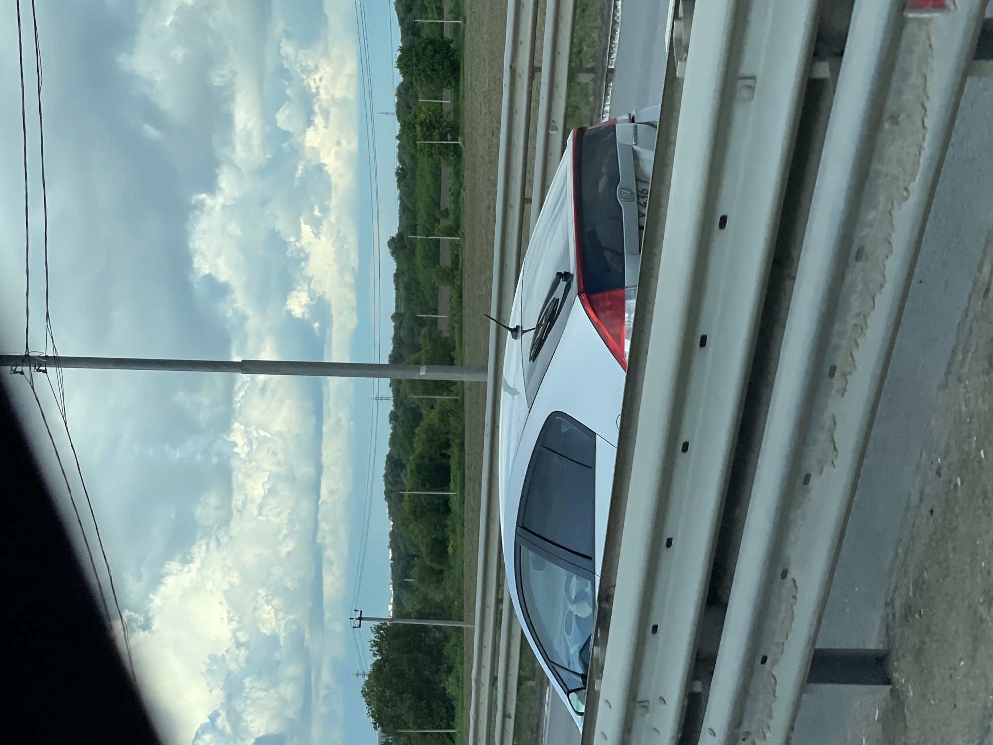 A white car is seen on a highway, as viewed from another vehicle. The scene is set against a backdrop of cloudy skies and a landscape of green fields and trees punctuated by utility poles. The car to the side appears to be a Toyota Prius, identifiable by its rear lights. A guardrail is visible between the viewer and the other car.
I'm unable to verify if this image is from Strelkovo, Russia, or if it's from Russia at all.
