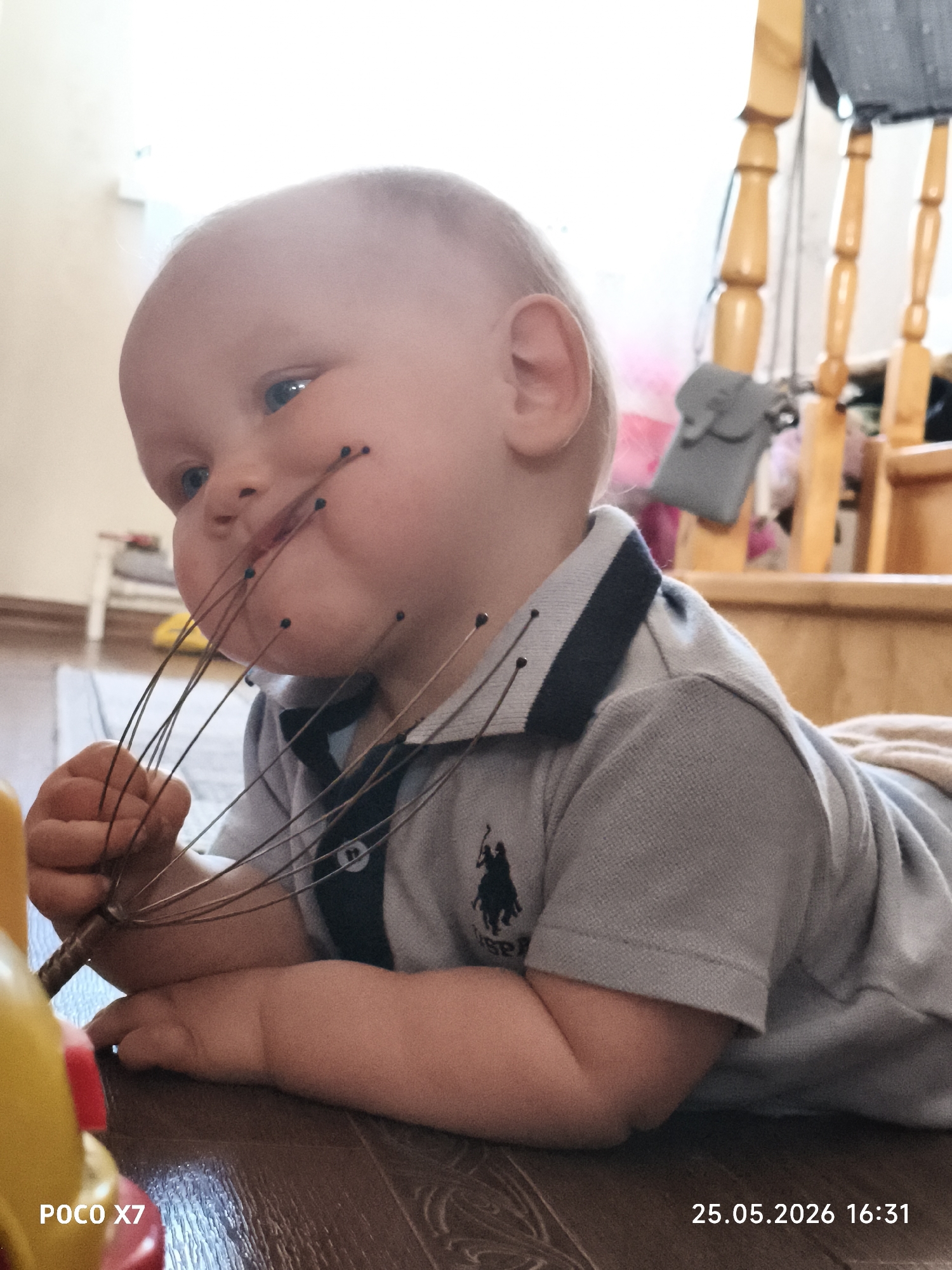 A young child is lying on the floor indoors, with a wooden head massager in their mouth. The child is wearing a gray polo shirt with a black logo on the left breast. The floor is brown wood, and there is a wooden staircase in the background. The date "25.05.2026 16:31" is stamped in the bottom right corner of the image. A yellow toy is visible in the bottom left corner. The location is an unidentified interior in the city of Unknown, Unknown.
