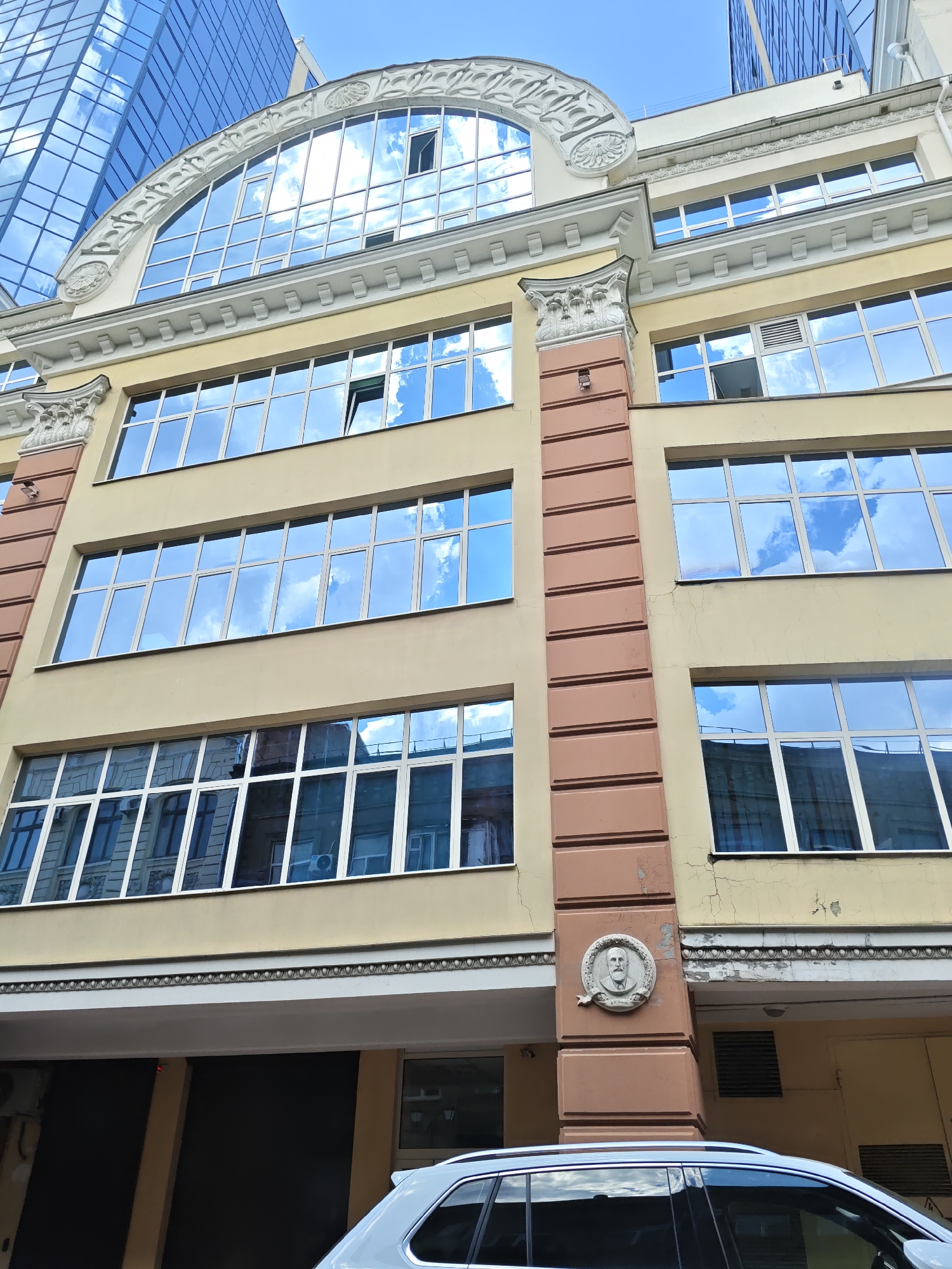 The image shows the facade of a building in Rostov-on-Don, Russia. The building has a pale yellow exterior and features multiple floors with large, reflective windows that mirror the blue sky and clouds. Architectural details like decorative moldings and pilasters are visible. A round relief sculpture of a man's head is mounted on the facade. In the foreground, the roof and rear window of a white car are visible. The context of Rostov-on-Don is provided by external information.
