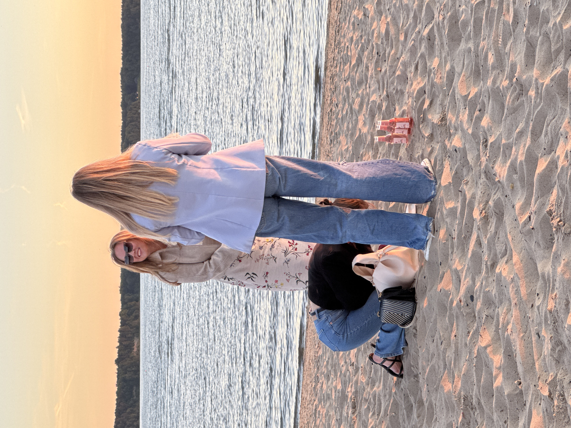 Three women are gathered on a sandy beach next to a body of water, possibly the Baltic Sea, at sunset. The sky is a warm orange and yellow, casting a glow on the scene. The woman on the left is wearing a beige trench coat and sunglasses, smiling at the woman in the middle. The woman in the middle is kneeling on the sand, facing away from the camera, with her head bowed. She is wearing a black top and blue jeans. A black purse and a light-colored tote bag are on the sand next to her. The woman on the right is standing, facing away from the camera, wearing a light blue blazer and wide-leg blue jeans. Three pink glass bottles are placed on the sand in front of her. The water is calm with gentle ripples. The background shows a dark treeline along the horizon. The overall impression is a casual, social gathering on the beach during golden hour. The location is the city of Tallinn, Estonia.