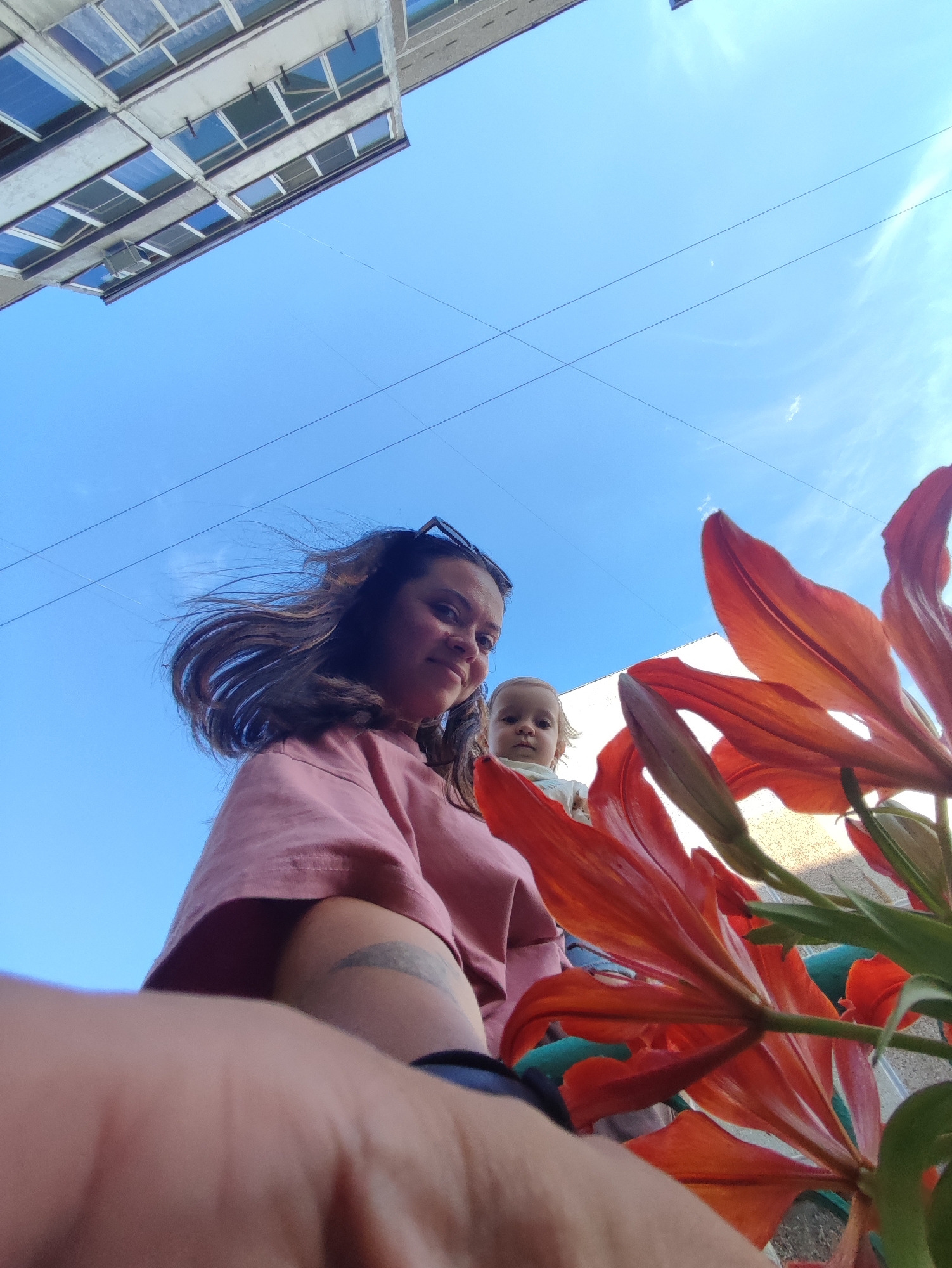 A low-angle, wide shot captures a woman holding a baby outdoors against a bright blue sky. The woman, with dark hair blowing in the wind, wears a pink t-shirt and is positioned on the left side of the frame. To her right, a baby with blonde hair looks towards the camera. In the foreground, bright orange lilies dominate the lower right portion of the image. In the background, a multi-story building with numerous windows and some power lines are visible. The scene is set in Chaikovsky, Russia.
