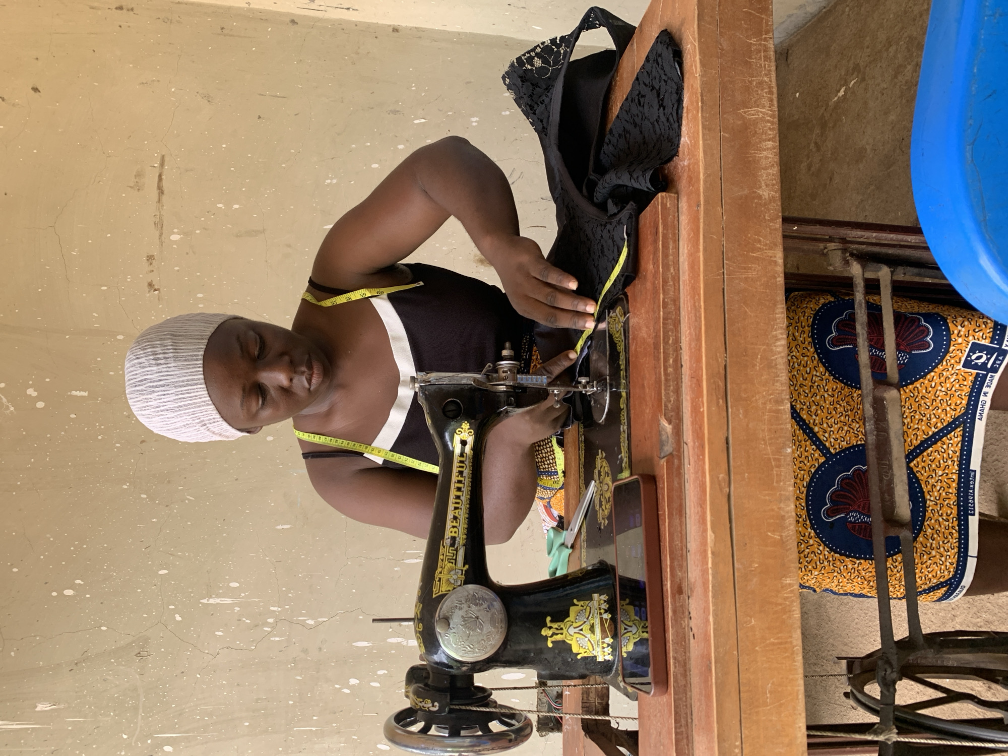 A person is sewing on a vintage black sewing machine. They are wearing a white and grey striped headscarf and a black tank top with white trim. A yellow measuring tape is draped around their neck. The sewing machine is on a wooden table, and a black fabric with lace detail is being sewn. A pair of green scissors lies on the table next to the sewing machine. A brown smartphone is positioned on a wooden block in front of the sewing machine. Behind the person, a wall is visible with white paint splatters. The person's lower body is covered by a brightly colored African print fabric skirt with a pattern of a bird and yellow and blue circles. The scene appears to be an indoor workspace or a covered outdoor area, possibly in Akwanga, Nigeria, given the context.