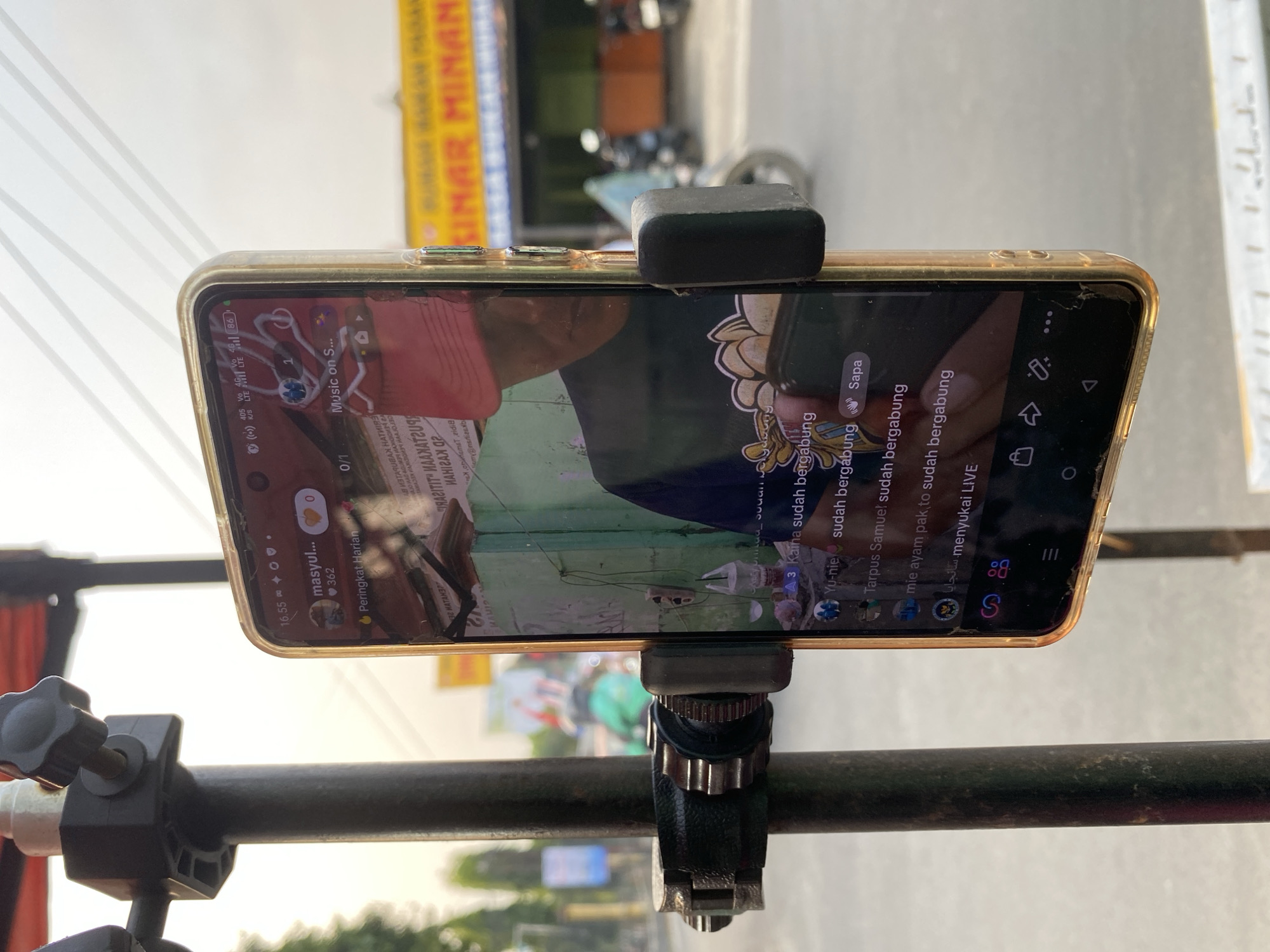 The image shows a smartphone mounted on a tripod in front of a building with a green wall. The smartphone screen displays a live streaming application, likely TikTok, with comments from viewers such as "Kaina sudah bergabung" and "Yu-nie sudah bergabung". The time displayed on the phone is 16:55, and the network shows 4G LTE. In the background, there is a street scene with a yellow banner that reads "SINAR MIMAN", suggesting the location is in Kasihan, Indonesia, a common area for businesses and commerce. The overall scene depicts a casual setting, possibly a roadside stall or shop, where the live stream is being broadcast.