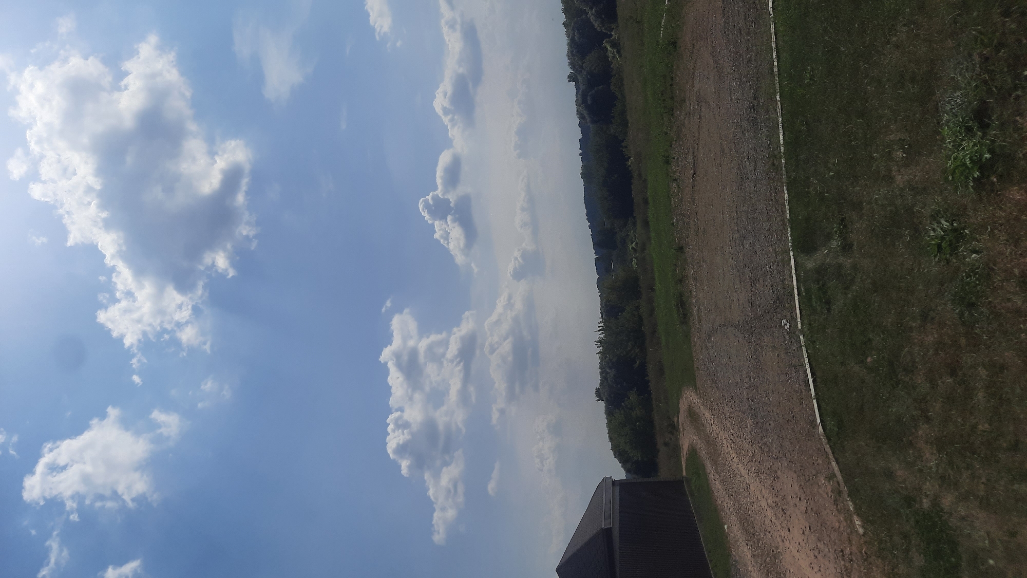 The image captures a wide view of a rural landscape under a partly cloudy sky. On the left side of the frame, a dark-colored, angular structure, possibly a building or shed with a dark, sloped roof and dark siding, occupies the foreground. Adjacent to this structure is a dirt or gravel path that curves away into the distance, leading towards a treeline. The path appears to be well-worn.
The middle ground features a flat expanse of grass and some dry, gravelly areas. Beyond this, a dense line of trees forms a natural boundary, with various shades of green indicating a mix of foliage. The background is dominated by a bright blue sky filled with scattered white and grey cumulus clouds. Sunlight filters through the clouds, creating subtle rays of light visible in the sky. The overall scene suggests a serene, undeveloped area, possibly on the outskirts of a town or in a more remote rural setting. The location is identified as Shklou, Belarus.