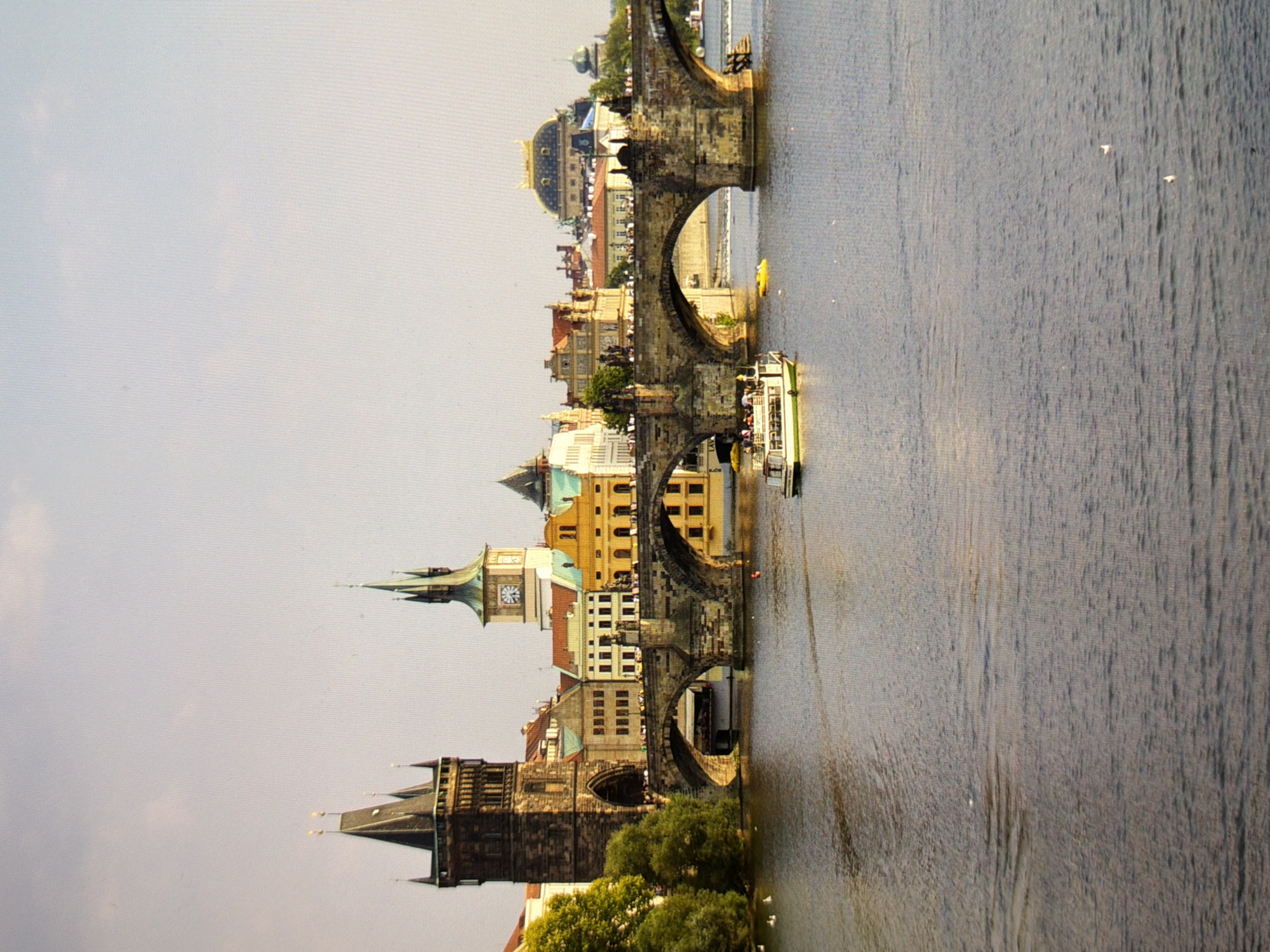 A vertical, eye-level, medium shot captures the Charles Bridge in Prague, Czech Republic, spanning the Vltava River under a hazy sky. The stone bridge, constructed of dark, weathered blocks, features multiple arches supporting a pedestrian walkway. On the bridge, figures are visible, appearing as small silhouettes. A tour boat with a white upper deck and green trim navigates the water to the right of the bridge, its passengers appearing as indistinct shapes. A small, bright yellow object, possibly another boat or marker, floats in the water near the bridge's right end.
Dominating the left side of the frame is a tall, dark stone tower with arched openings and spires. Behind it, various buildings with red-tiled roofs and colorful facades, including a prominent building with a clock tower, are visible. The water of the Vltava River occupies the lower half of the image, reflecting the soft, overcast light. The water's surface is rippled, with a few small birds visible on the water towards the bottom left. The overall lighting suggests it is daytime, possibly late afternoon.