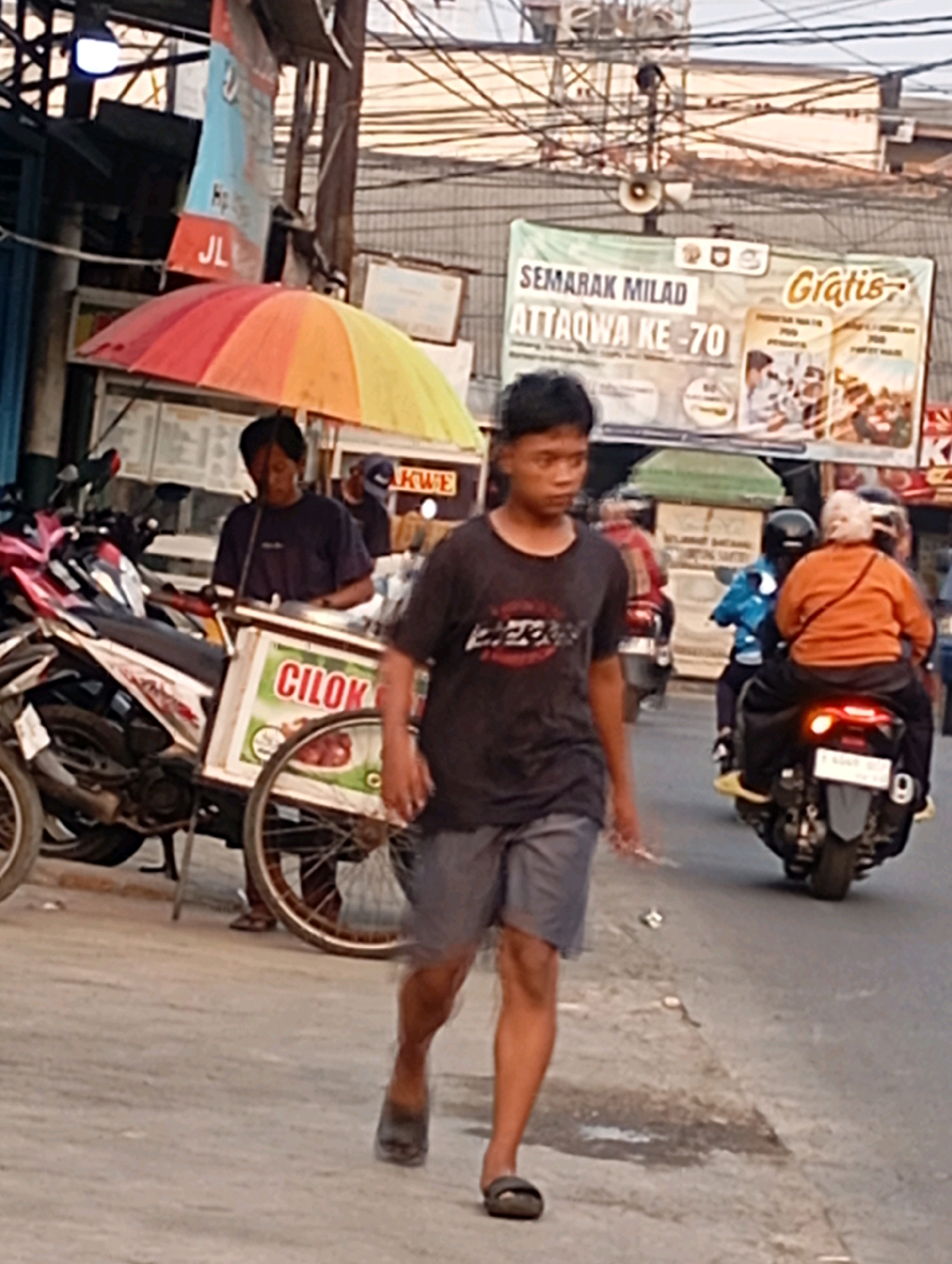 In Babelan, Indonesia, a street scene unfolds with a young man walking towards the viewer. He is wearing a dark t-shirt and grey shorts, with dark sandals on his feet. To his left, a vendor is preparing food from a cart that is decorated with a sign that reads "CILOK." Behind the vendor, motorcycles are parked. A colorful umbrella is positioned over the vendor's cart. In the background, banners with Indonesian text are visible, and several people are riding motorcycles on the street. The environment appears to be an urban setting with overhead wires crisscrossing the sky.