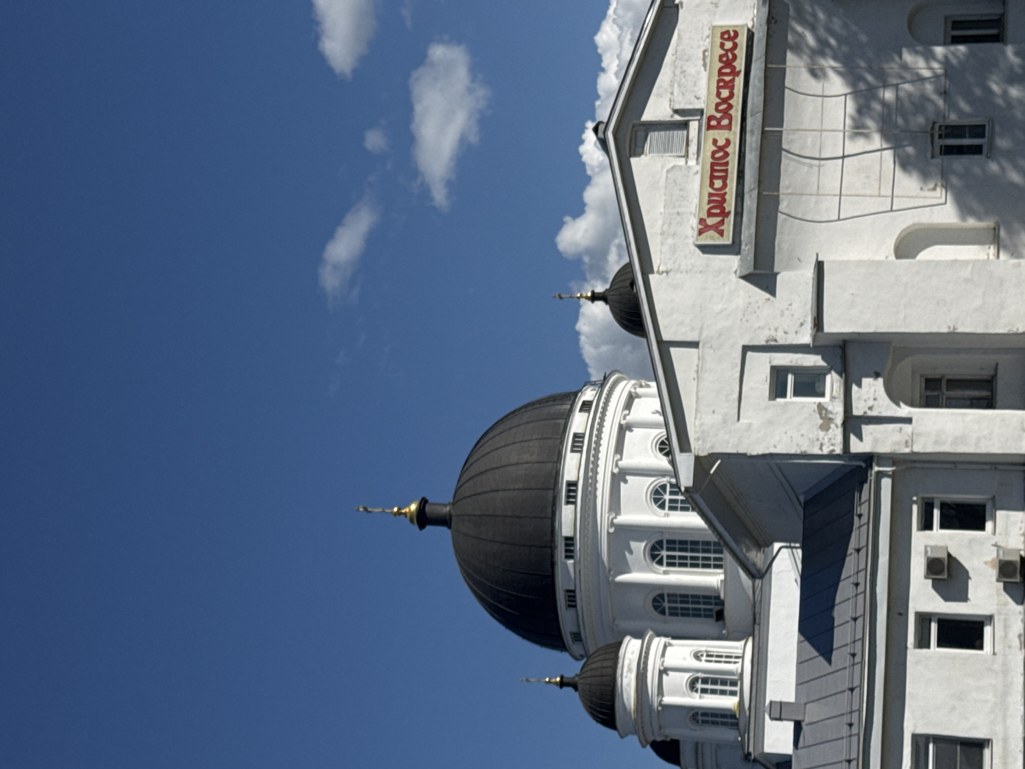 This image shows a close-up view of a white, domed cathedral with a dark dome, under a bright blue sky with scattered clouds. The cathedral has multiple white structures with windows and smaller domes. A red sign with yellow lettering is visible on one of the white structures. The sign appears to read "Христос Воскресе," which translates to "Christ is Risen" in English. This cathedral is likely the Alexander Nevsky Cathedral, a prominent landmark in Nizhny Novgorod, Russia, known for its Neo-Byzantine architecture and distinctive white facade. The scene is bathed in natural sunlight, casting shadows on the building. No people are visible in the image.