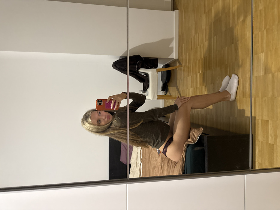 A mirror selfie captures a slender, blonde woman in a long-sleeved brown top and dark underwear, seated on a bed. Her legs extend to the right, clad in white socks. Behind her, a simple white chair holds dark clothing. The background includes white walls and wooden floors, reflecting the bedroom scene.