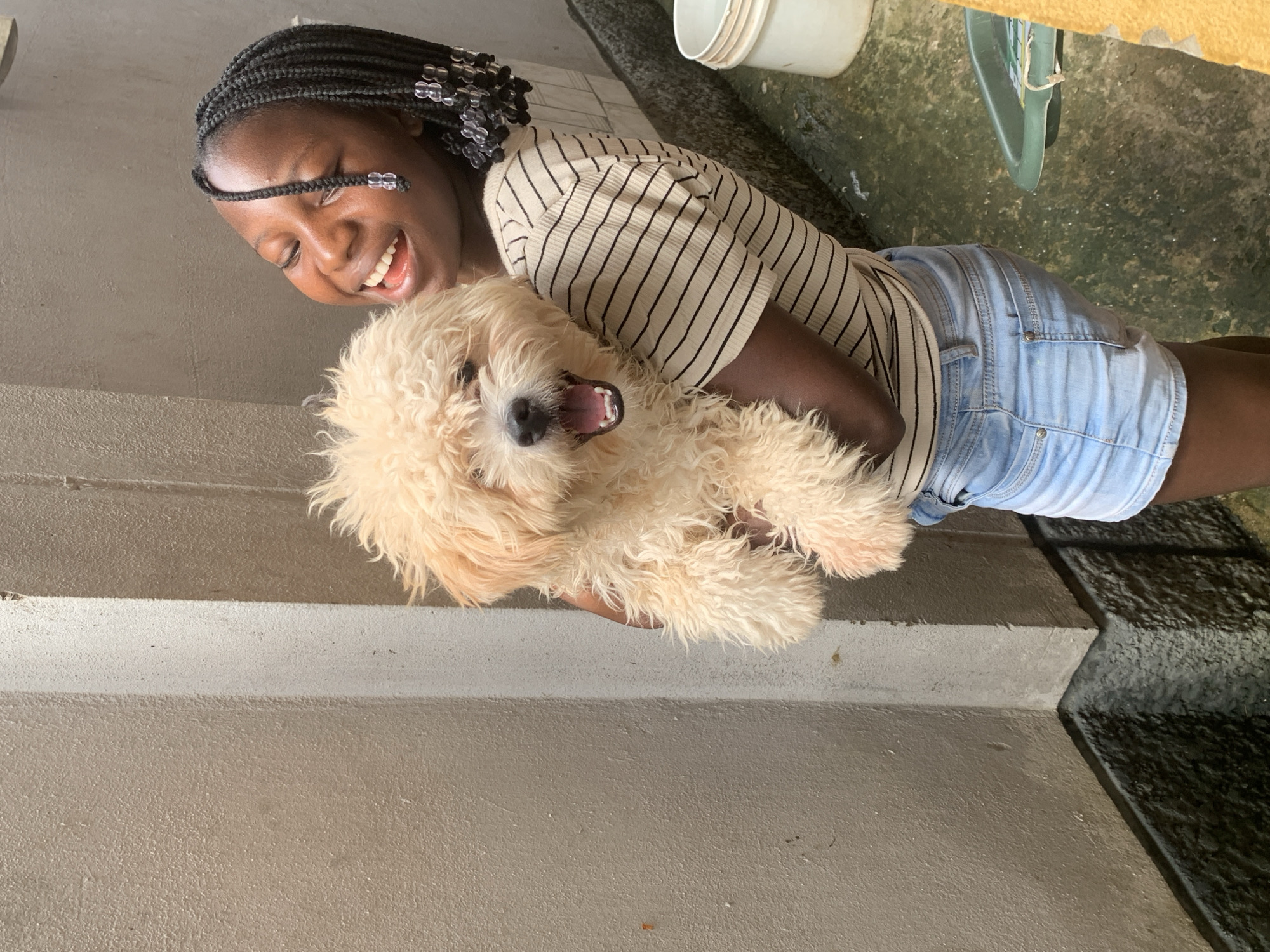 A young Black girl, with braided hair decorated with transparent beads, is holding a light-colored, fluffy dog. The girl is wearing a striped t-shirt and denim shorts and is smiling broadly, with her eyes closed. The dog, which appears to be a poodle mix, is looking at the camera with its mouth open. They are standing outdoors next to a beige and gray concrete wall. The ground appears to be wet and there is a white bucket and a green plastic object visible in the background. The scene is outdoors, likely in a residential area of Rido, Nigeria.