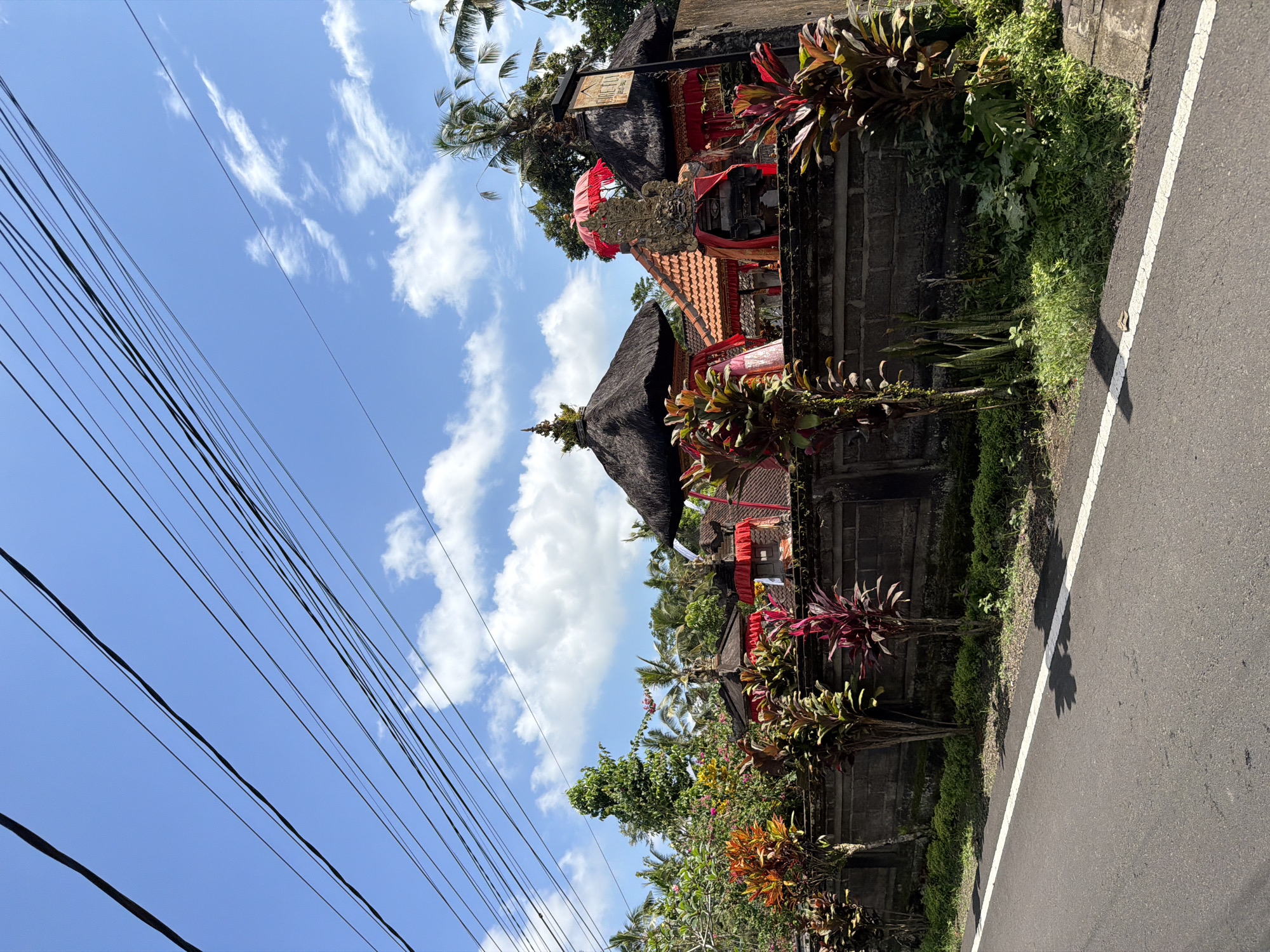 This is a medium shot, taken from a low angle, of a street scene in Tampaksiring, Indonesia. The sky is a bright blue with scattered white clouds and a tangle of black electrical wires stretched across the top of the frame. A paved road with a white line down the center occupies the bottom third of the image. To the right of the road is a low, dark brick wall that runs horizontally across the frame, topped with lush green vegetation and vibrant tropical plants with red and green leaves. Behind the wall, a traditional Balinese compound is visible. The compound features several multi-tiered, thatched-roof structures, adorned with red fabric and ornate stone carvings. A prominent, intricately carved stone sculpture is centrally positioned, topped with a bright red umbrella. A wooden sign with "ORE FLY" written in yellow lettering hangs on a post to the right of the compound. The scene is bathed in bright sunlight, casting shadows on the road and highlighting the colors of the vegetation and the compound decorations.