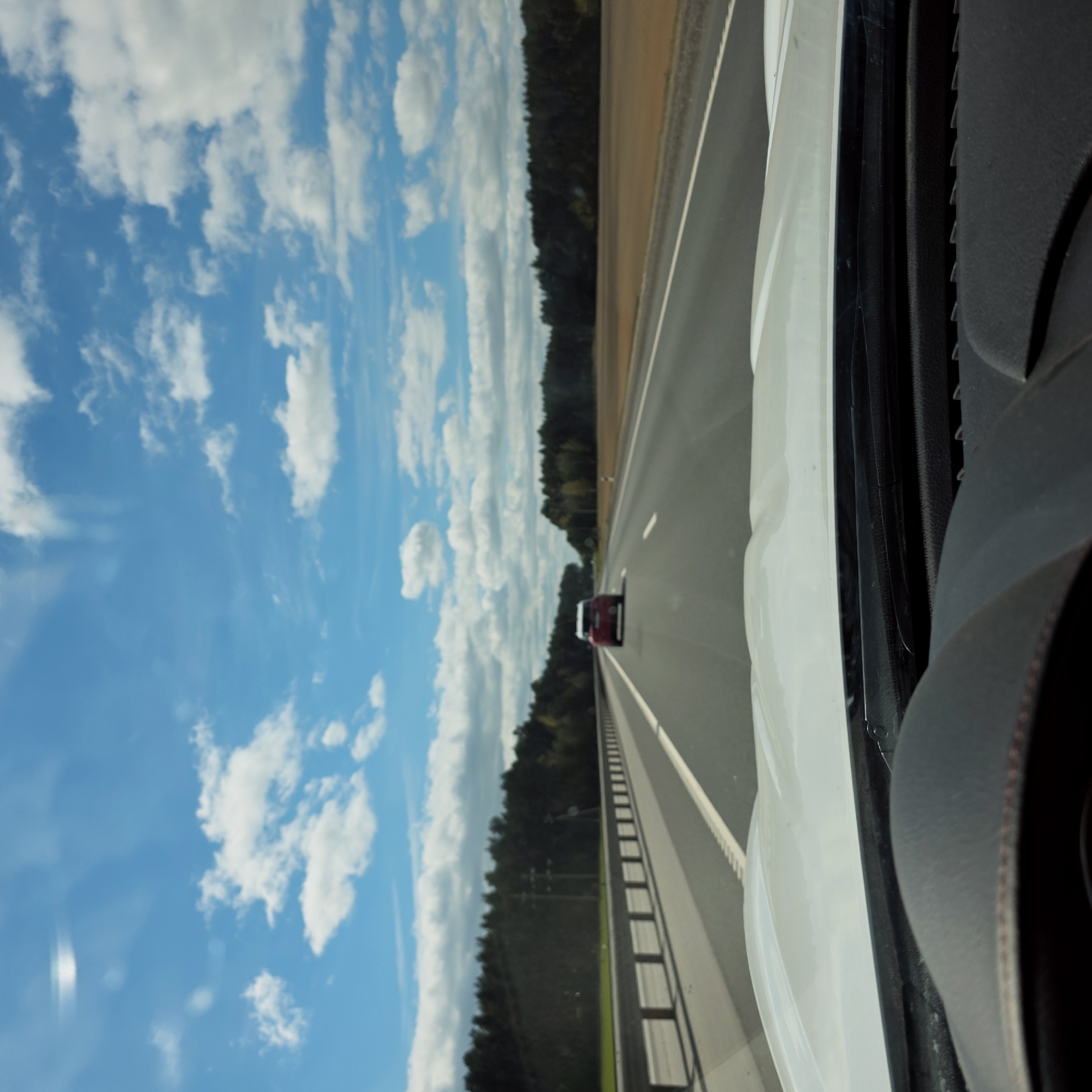 This image captures a bright daytime scene from inside a moving vehicle on a highway in Скідзельскі сельскі Савет, Belarus. The perspective is from the passenger side, looking forward and to the left through the window.
On the right side of the frame, parts of the white vehicle's exterior and a dark grey interior element, possibly a dashboard or door panel with textured surfaces, are visible. The main focus is the open road ahead. A multi-lane asphalt highway, delineated by clear white lane markings, stretches into the distance. A metal guardrail runs along the left edge of the road. Further down the highway, a red car is seen, traveling in the same direction.
Beyond the road, a dense, dark green forest forms a consistent tree line along the horizon, suggesting a rural or semi-rural setting. A strip of lighter brown earth or shoulder runs between the highway and the forest. Above, the sky is a vibrant blue, adorned with numerous fluffy white cumulus clouds, indicative of clear and fair weather. Some minor reflections or lens flares are present in the upper sky, likely from the vehicle's window. The overall impression is one of a calm journey through a forested landscape under a sunny sky. No visible text is present in the image.