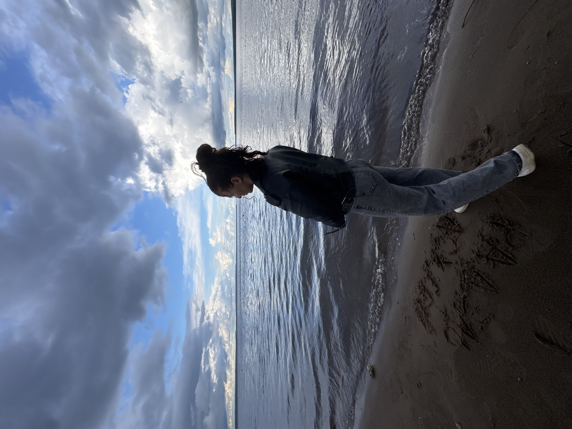 The image depicts a person standing by the edge of a large body of water with a sandy or pebbly shoreline. The scene is set in Chaikovsky, Russia, likely along the Kama River or Votkinsk Reservoir.
The main subject is a person, seen in profile, with dark hair tied up in a messy bun. They are wearing a dark jacket (possibly leather), light-wash blue jeans, and white sneakers. The person is looking downwards, towards the water and sand, suggesting a moment of contemplation or observation.
The sky above is partially cloudy, featuring a mix of dramatic dark grey clouds, lighter white clouds, and patches of bright blue sky. This indicates daytime, possibly a late afternoon or early evening setting given the lighting. The expansive water surface is textured with ripples and reflects the overhead sky. Gentle waves break on the shore, creating lines of white foam along the waterline. The sand on the beach is dark and appears wet near the water's edge.
A notable detail is the word "KAMA" visibly written in large letters on the sand in the foreground, near the person's feet. Other faint, indistinct markings are also present on the sand. The overall mood conveyed is one of quiet solitude and reflection amidst a natural landscape.