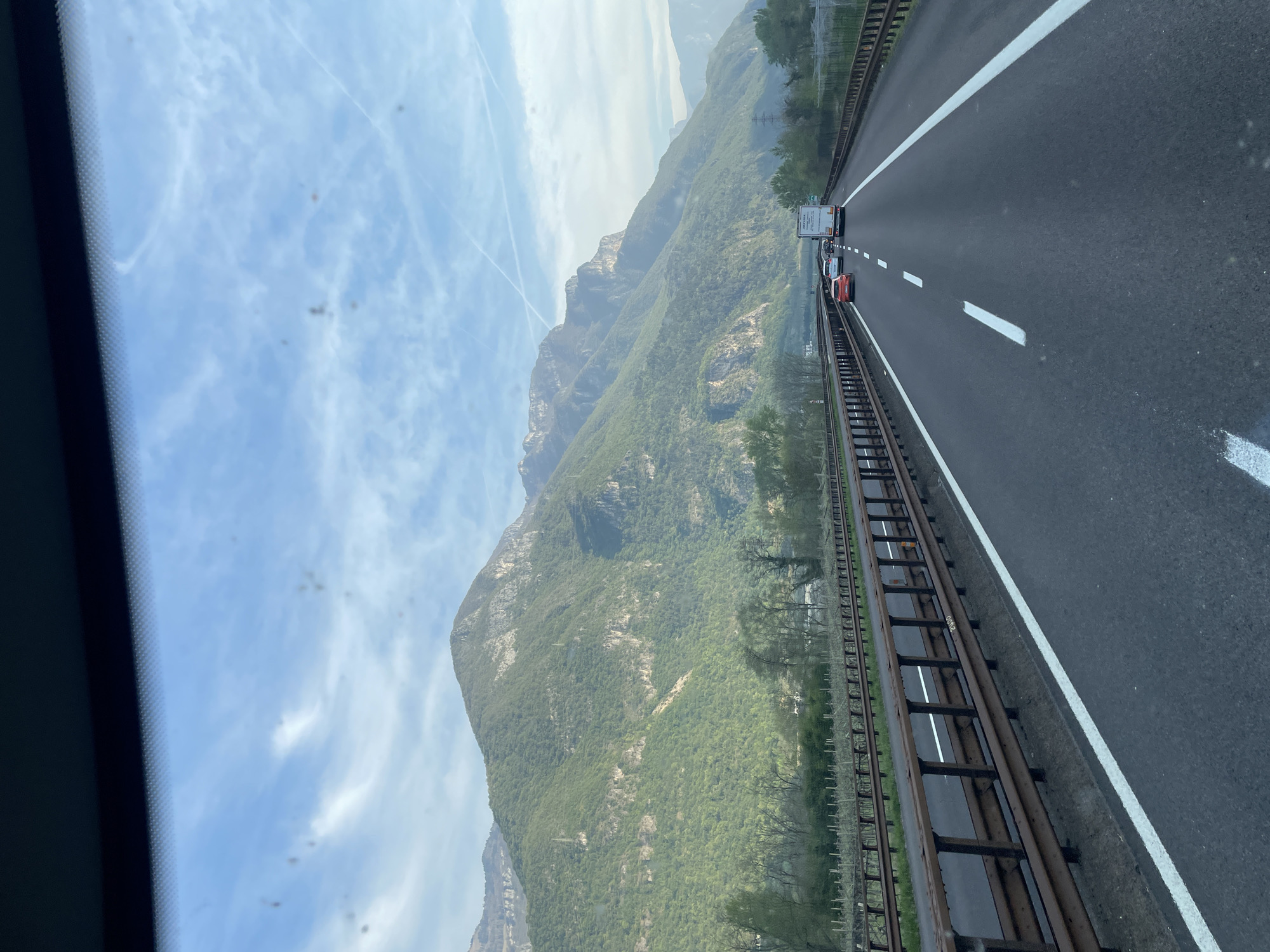 The image shows a view from inside a vehicle, likely a car, driving on a highway. The location appears to be near Dolcè, Italy, judging by the presence of mountains and the type of road. The road is a multi-lane highway with white lane markings and a metal guardrail separating the lanes from a hillside covered with greenery.
There are several vehicles on the road, including what appears to be a red car and a box truck.
The background features a mountain range partially obscured by haze. The sky is blue with scattered white clouds and contrails. The scene conveys a sense of travel and the natural landscape of the region.