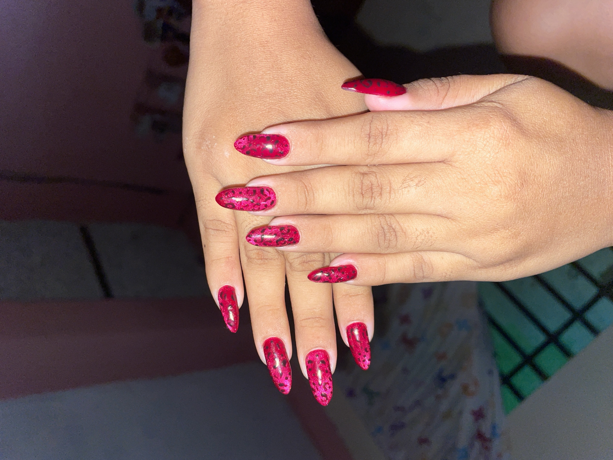 The image shows a pair of hands with manicured fingernails. The fingernails are long, almond-shaped, and painted with a bright red polish featuring a darker red speckled design. The hands appear to be resting on a surface. The background is indistinct, showing blurred shapes and colors. The location and specific environment are unknown.