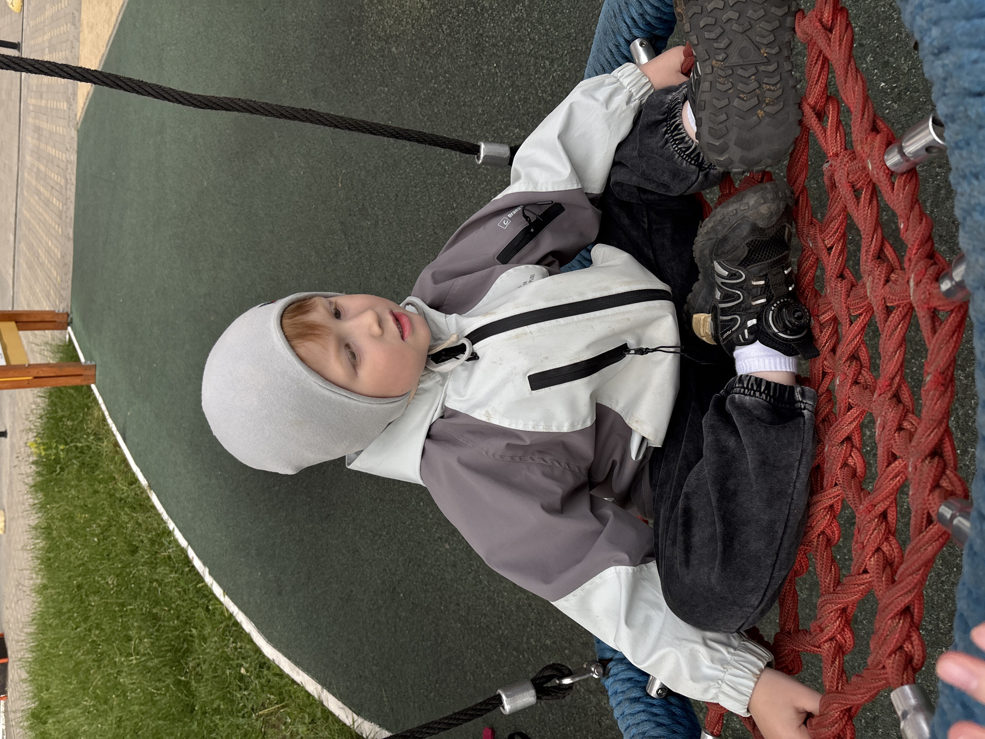 A young boy, approximately 4-7 years old, with reddish-brown hair partially visible, is seated centrally in a circular nest swing. He wears a light gray beanie, a two-tone jacket primarily light gray/off-white with gray shoulders and sleeves, dark gray pants with elasticized cuffs, white socks, and black athletic shoes with textured soles. The jacket shows light dirt marks, and a small logo is visible on the right sleeve pocket. The boy's gaze is directed towards the right side of the frame.
The swing's seat is constructed from thick, interwoven red ropes forming a mesh, secured by metal components and suspended by dark ropes, two of which are visible extending upwards. A section of blue rope, part of the swing's structure, is visible on the left side.
The ground beneath and around the swing is a dark green rubberized safety surface, characteristic of playgrounds. This surface is bordered by a distinct curved white line, beyond which is a strip of well-maintained green grass. In the background, past the grass, a paved area with light brown interlocked pavers is visible, along with parts of wooden playground structures.
The scene depicts an outdoor playground setting. Due to the generic nature of the playground equipment and environment, the specific location within the city of Izhevsk, Russia, cannot be precisely determined from the provided image.