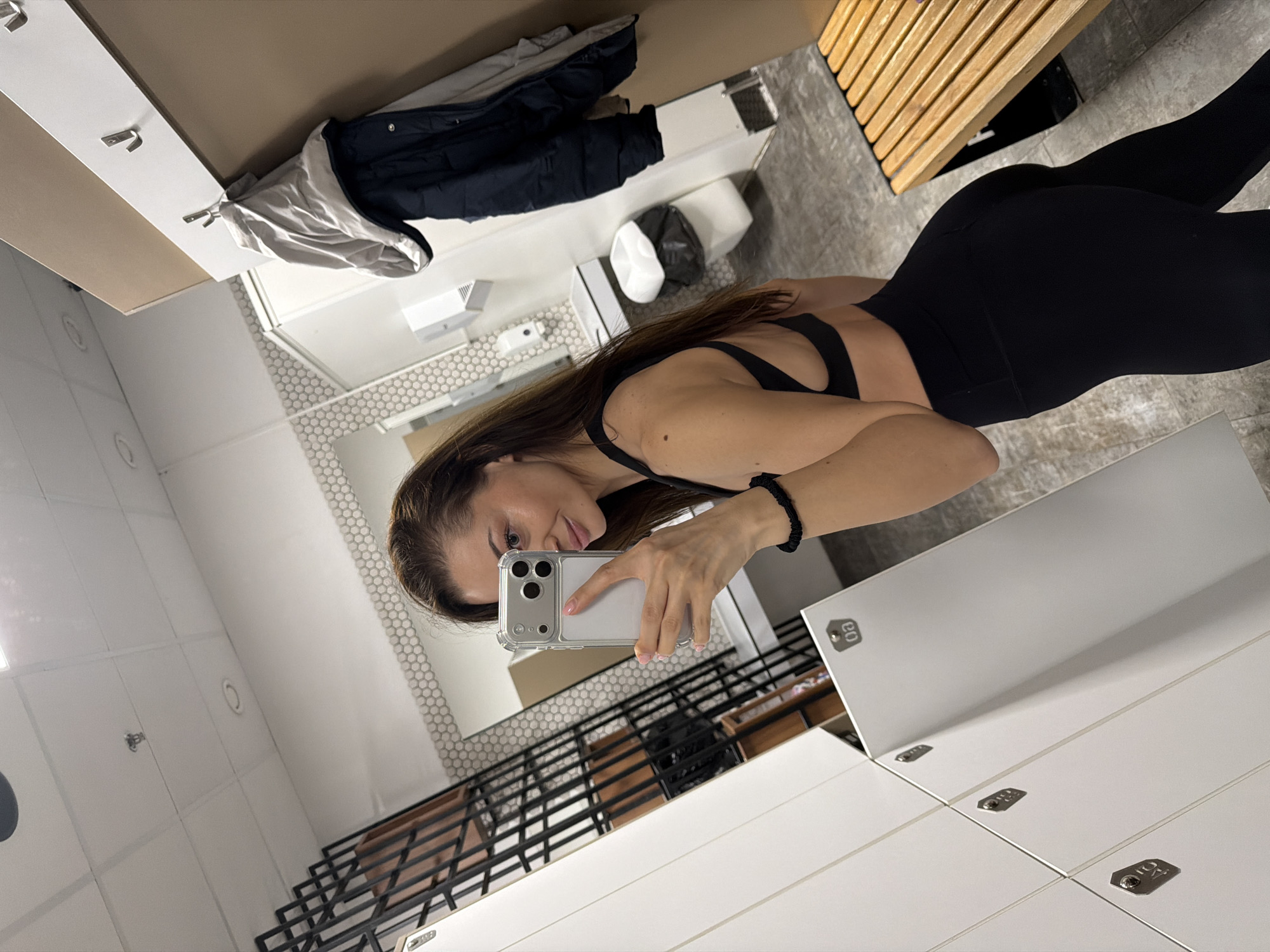 The image depicts a woman in a gym locker room. She is taking a selfie with her smartphone, wearing a black sports bra and leggings. The locker room is equipped with white lockers and wooden benches. A mirror on the wall reflects the bathroom area, including a sink and toilet. Clothing items, such as jackets, are hanging on hooks near the lockers. The location is likely in Varna, Bulgaria, due to the implied gym setting and the user's request.