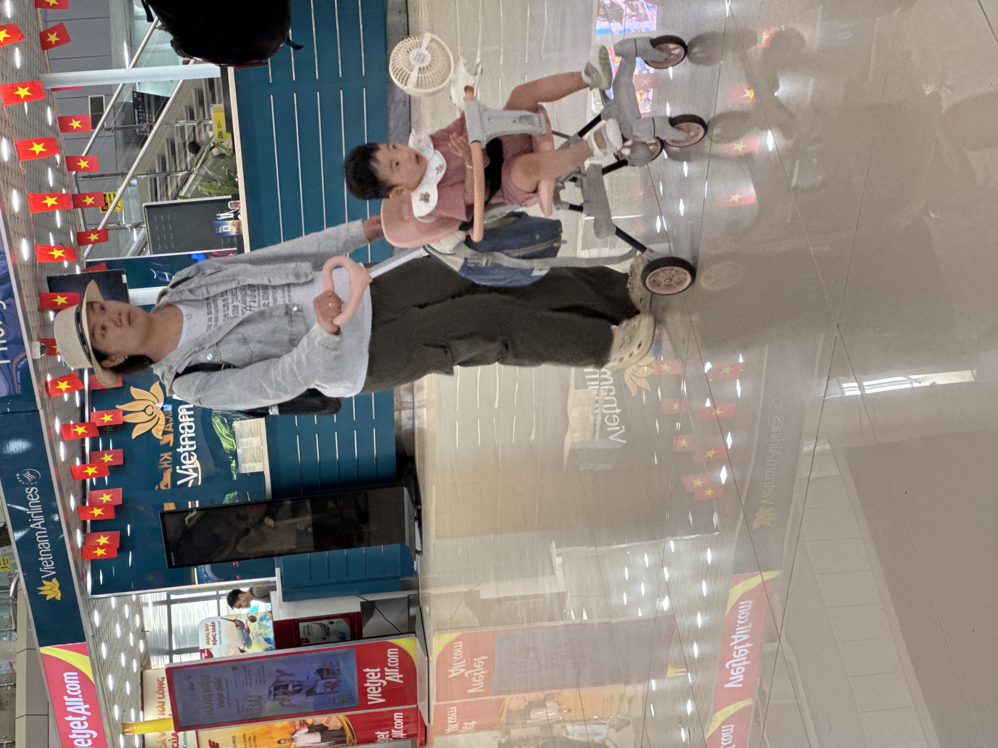 The image shows a woman pushing a stroller with a child in it, in what appears to be an airport terminal. The woman is wearing a straw hat, a denim jacket, and dark pants. The child is wearing a pink outfit and is looking forward with their mouth open. The stroller is pink and grey. In the background, there are signs for Vietnam Airlines and Vietjet Air. There are also Vietnamese flags hanging from the ceiling. The floor is tiled and reflective, showing the reflections of the signs and flags. The scene appears to be in the city of Bac Cam Ranh Ward, Vietnam, based on the signage.