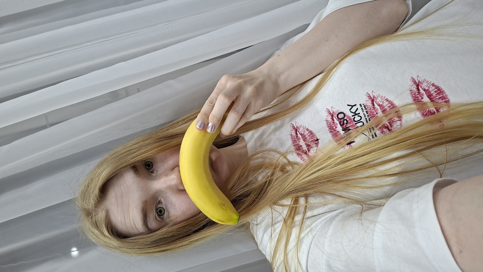 A woman with long blonde hair lies on a white surface, possibly a bed, in a location that appears to be inside a room with white curtains. She holds a yellow banana near her face. She wears a white t-shirt printed with red lipstick marks and the words "Lucky". The woman has fair skin and green eyes. Her fingernails are painted with a silver finish. The image does not explicitly depict the city of Chaikovsky, Russia.