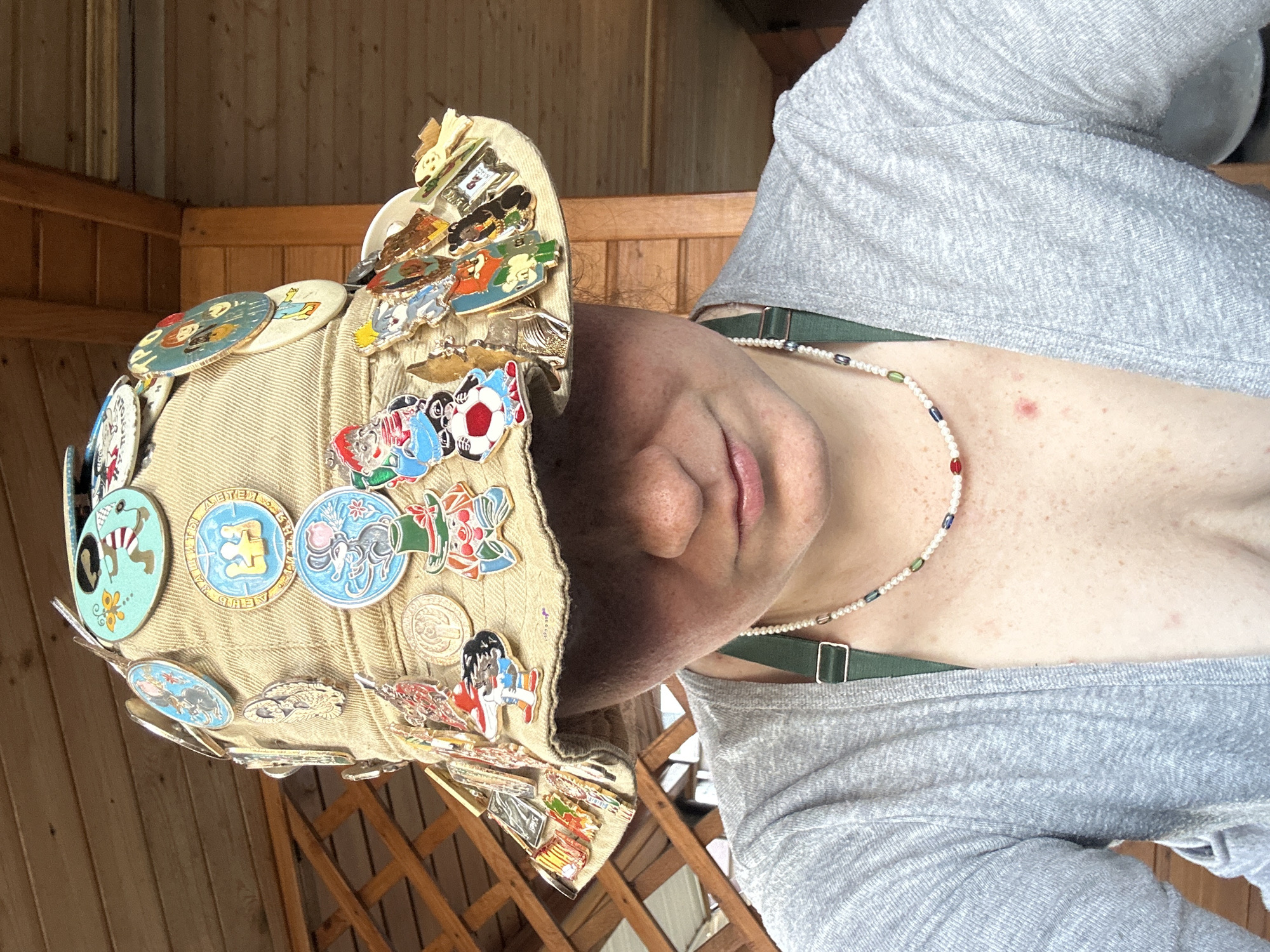A person wearing a beige hat adorned with numerous pins and buttons is photographed in what appears to be an outdoor setting, possibly a porch or patio, suggested by the wooden walls and lattice in the background. The person is wearing a gray shirt and a delicate pearl necklace with colored beads. The subject's face is partially obscured by the hat, with only the nose and mouth visible. The camera angle is slightly tilted, and the person seems to be looking directly at the camera. There is no clear indication of the location being in Nizhny Novgorod, Russia.