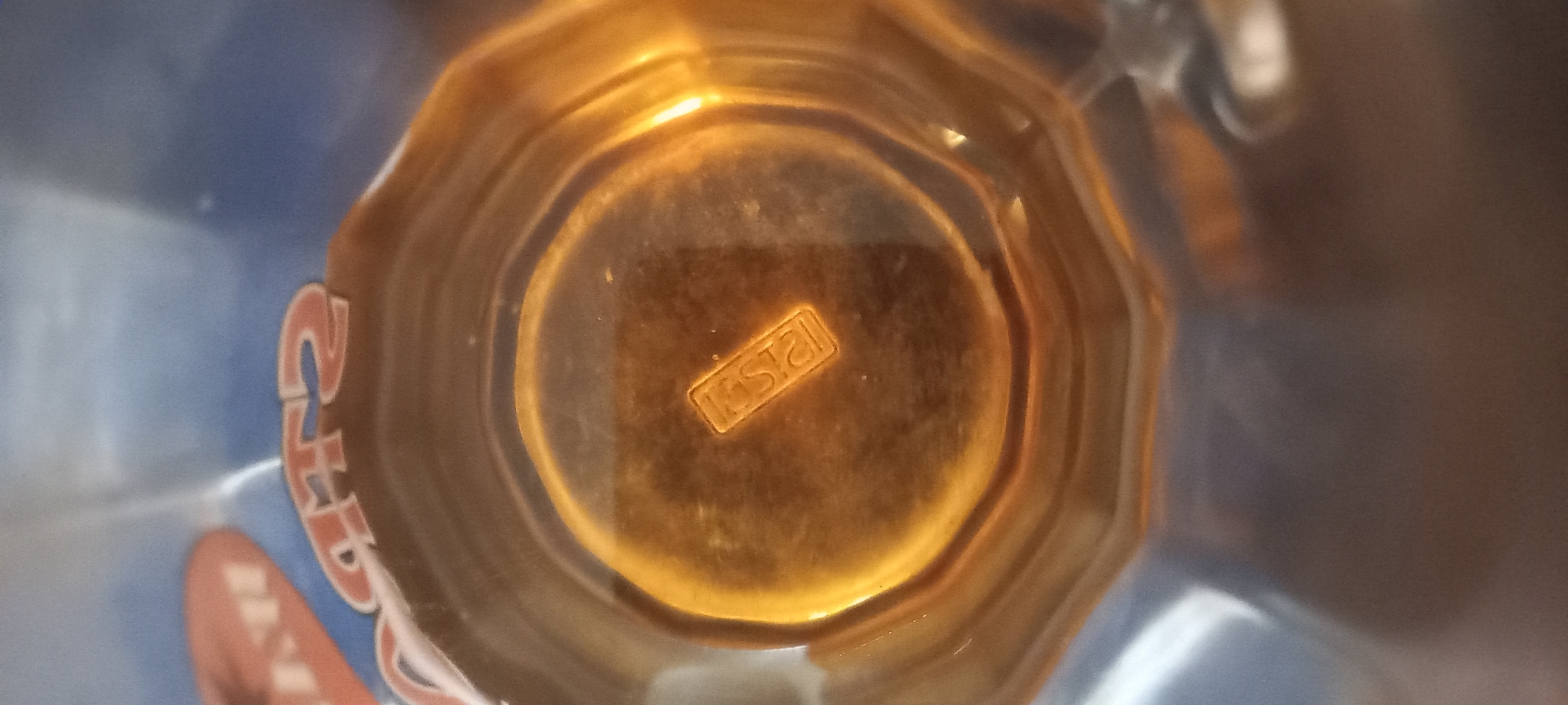The image is a close-up, overhead view of the bottom of a glass or plastic container. In the center of the base, there is a raised rectangular emblem with the word "KRYSTAL" embossed in capital letters. The rest of the base has a faceted, octagonal shape. The material of the container appears to be tinted a warm amber color due to the lighting and reflection. The background is out of focus and appears to be a blue surface with some blurred red lettering, possibly from a label or packaging. No people or specific location details within Kryvyi Rih, Ukraine are visible.