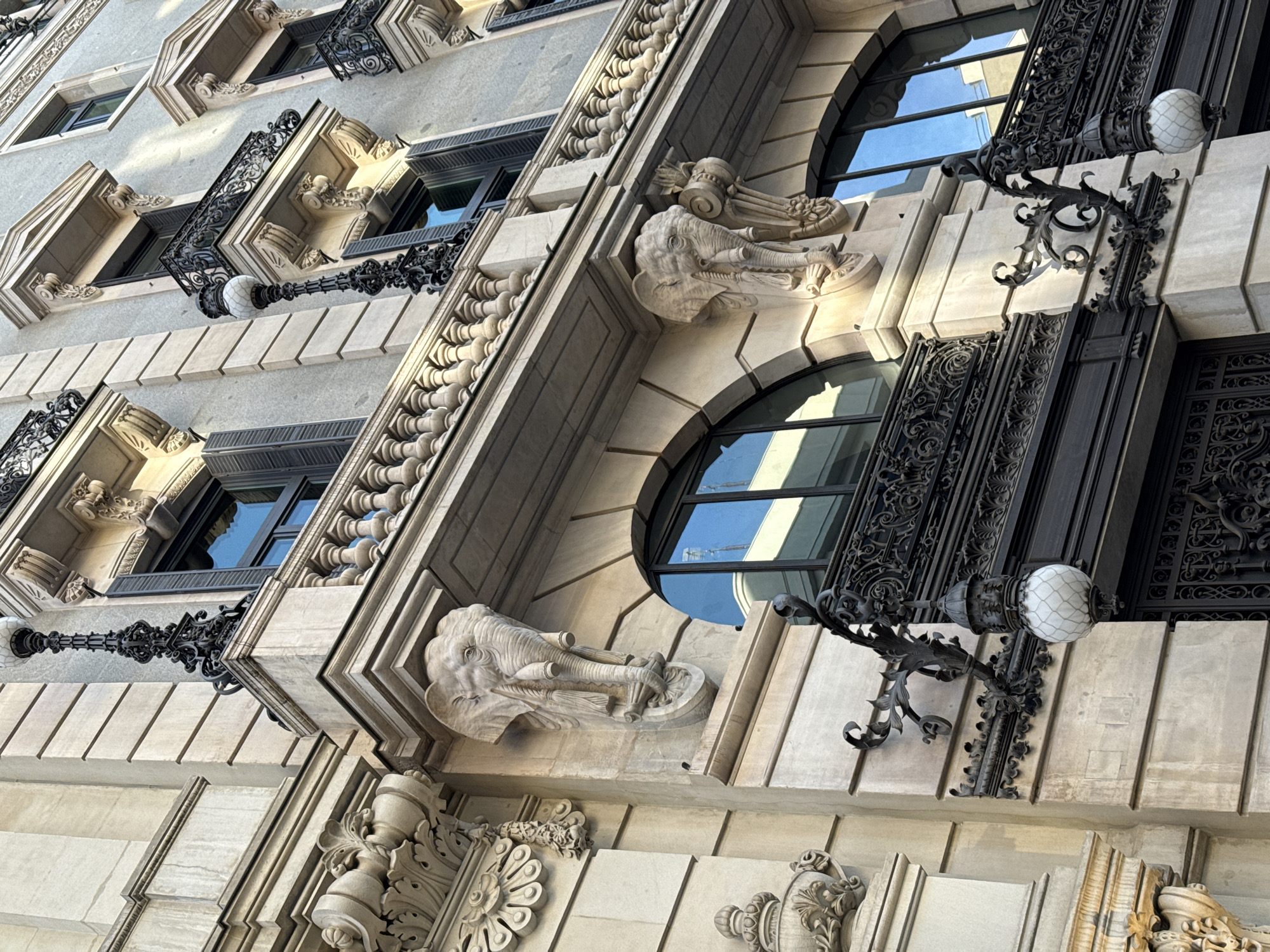 The image showcases the architectural details of a building facade, likely located in Madrid, Spain. The style appears to be historic, with elements of Beaux-Arts or similar classical revival styles.
Specific details include:
* **Windows:** A mix of arched and rectangular windows are visible, some framed by ornate wrought iron balconies or grilles.
* **Ornamentation:** Elaborate stone carvings and decorative elements adorn the facade. These include sculpted elephant heads, floral motifs, and other classical details.
* **Lighting:** Decorative street lamps, with globe-shaped diffusers, are attached to the building's exterior.
* **Materials:** The building is constructed of stone, likely limestone or a similar material, with wrought iron details.
* **Perspective:** The image is taken from a low angle, looking upwards at the building.
The overall scene portrays a sense of historical grandeur and architectural richness, characteristic of buildings found in Madrid's city center.