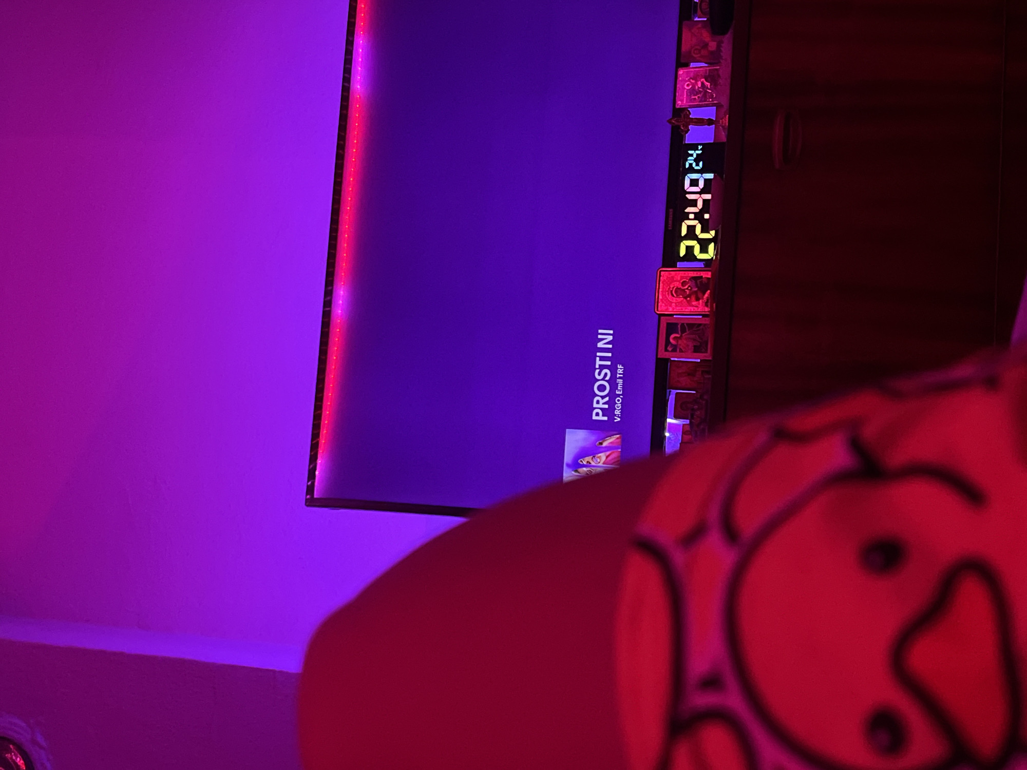 This image captures an indoor scene, likely in Debelt, Bulgaria, under strong purple ambient lighting. In the foreground, out of focus, a human leg or foot is visible, wearing a red slipper featuring a black-outlined cartoon duck face. In the mid-ground, a television screen displays the text "PROSTINI" and "V:RGO, Emil TRF" in white against a dark purple background. The top edge of the television screen is illuminated by a strip of red LEDs. Positioned below the television, on a dark furniture surface, a digital clock with multi-colored digits displays "22:49" and "24.". Several small framed pictures or religious icons are arranged on the surface around the digital clock. The environment suggests a relaxed, nocturnal setting.