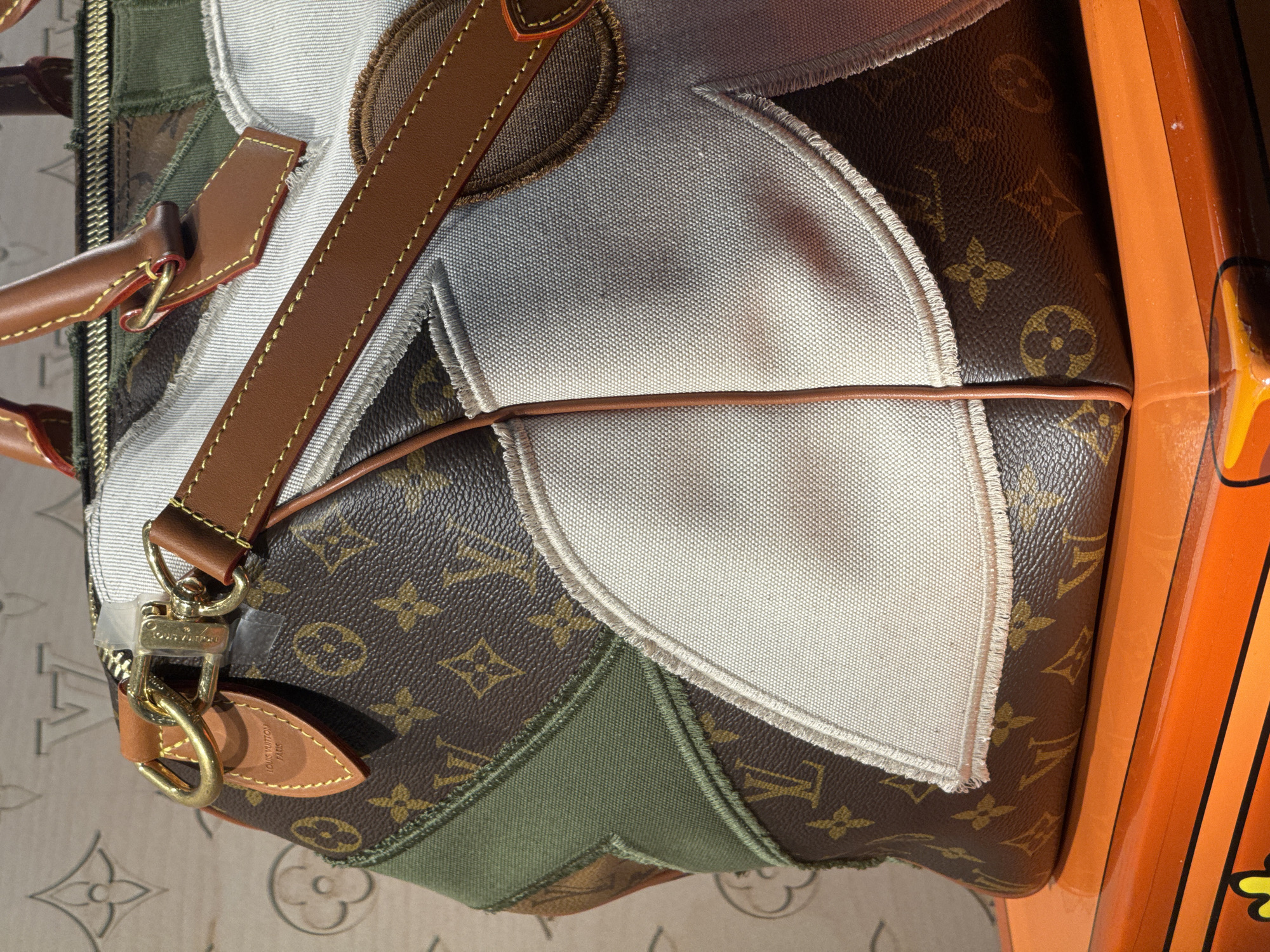 A close-up, eye-level shot captures a Louis Vuitton Speedy bag with a patchwork design. The bag is made of the iconic brown Louis Vuitton monogram canvas, interspersed with panels of white canvas, olive green fabric, and brown denim. The bag features brown leather handles and trim, with gold-toned hardware. A Louis Vuitton tag hangs from one of the handles. The bag rests on an orange surface with yellow accents, possibly a piece of furniture. The background appears to be a patterned surface, possibly wallpaper, with a faint Louis Vuitton motif. The image is well-lit, with the light source coming from the top right. The scene is indoors, with no discernible city or specific location information available within the image itself.