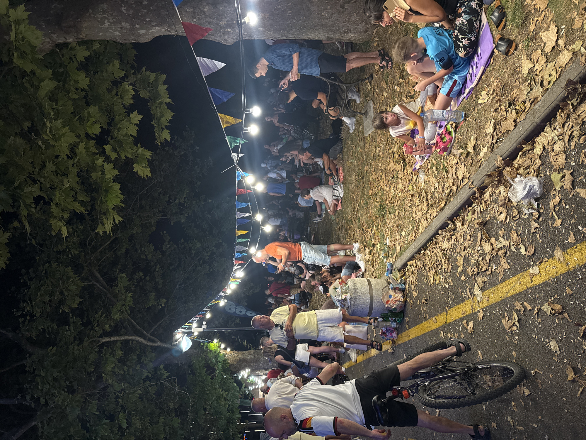 This image depicts an outdoor gathering at night, possibly a festival or event, in Burgas, Bulgaria, under a canopy of large trees. String lights and colorful triangular flags are strung between the trees, illuminating the scene.
In the foreground, a man is seated on a bicycle, facing away from the camera. To his right, on the asphalt road marked with a yellow line, are scattered fallen leaves, some plastic bottles, and a large concrete cylindrical object with trash on top.
Further into the scene, a diverse group of people are seen sitting and standing on the grass and pavement. Many are casually dressed, suggesting a relaxed atmosphere. Some individuals are engaged in conversation, while others are looking at their phones or listening to music.
In the mid-ground, a cluster of people are seated on blankets on the grass, including two children. One child is holding a pink water bottle and appears to be speaking. Another child, wearing a blue shirt, is sitting next to a partially consumed bottle of water. A woman is seated beside them, speaking on a mobile phone.
In the background, more people are visible, some standing and others sitting, with the light from the string lights creating a festive ambiance. The overall impression is one of a community event taking place in a park-like setting within the city.