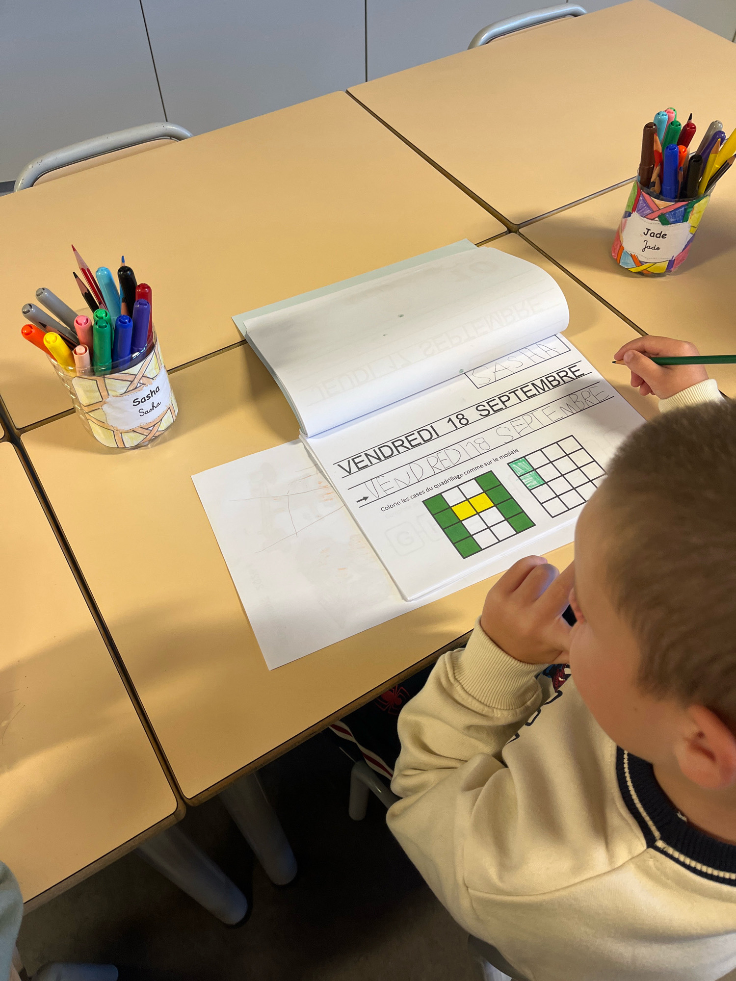 A young boy, viewed from over his shoulder, is seated at a light brown table in a classroom. He is wearing a cream-colored sweatshirt with a navy and white striped collar and is holding a green pencil in his right hand. In front of him is an open book displaying a grid with some squares colored green and one yellow square. The book has text that reads "VENDREDI 18 SEPTEMBRE" and further instructions in French. To the left of the book is a clear plastic cup filled with colored markers and pencils, with a label that says "Sasha Sasha". To the right of the book is another similar cup with markers and pencils, and a label that reads "Jade Jade". The table surface is a light beige, with visible seams indicating it is made of multiple sections. The background shows parts of a grey wall and a metal chair leg. This scene is consistent with a classroom setting, potentially in the city of La Fouillouse, France, given the French text on the book and labels.