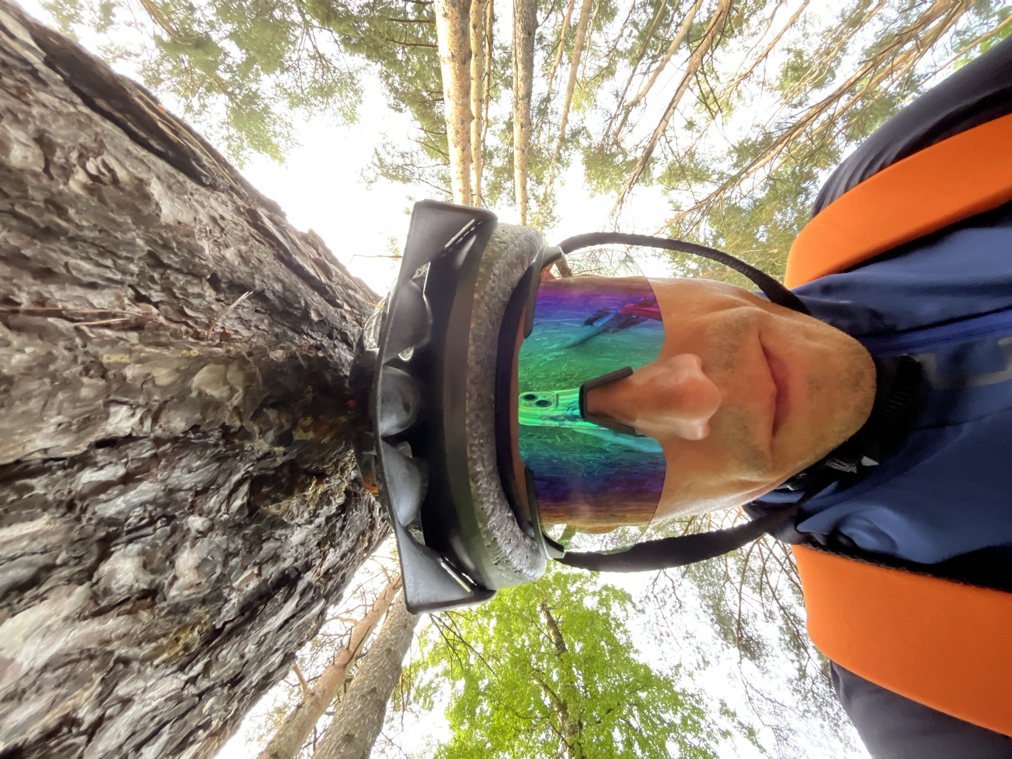 A selfie taken from a low angle looking upwards shows a man wearing a helmet with reflective goggles standing next to a tree. The man is wearing a blue jacket with orange straps. The background shows a forest of tall trees with green leaves. The location is likely Chaikovsky, Russia.