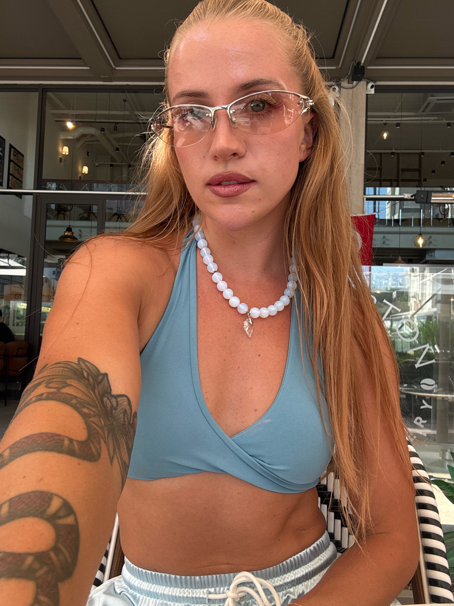 A young woman with blonde hair pulled back is sitting at a table in an outdoor cafe in Antalya, Turkey. She is wearing a blue bikini top, light blue shorts, and pink-tinted glasses. A beaded necklace with a pendant hangs around her neck. She has a tattoo of a snake on her left arm. The cafe appears to be modern with large glass windows and visible light fixtures. The background shows reflections in the windows, suggesting an indoor space. The woman is looking directly at the camera.
