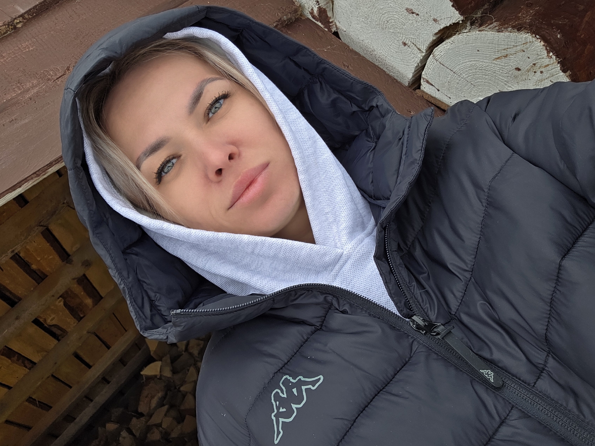 A woman with light blonde hair and blue eyes poses for a selfie. She is wearing a black puffer jacket over a white hoodie, with the hood of the jacket pulled up. The jacket has a small Kappa logo on the lower left side. The background appears to be a wooden structure or wall. The photo was taken in the city of Chaikovsky, Russia.