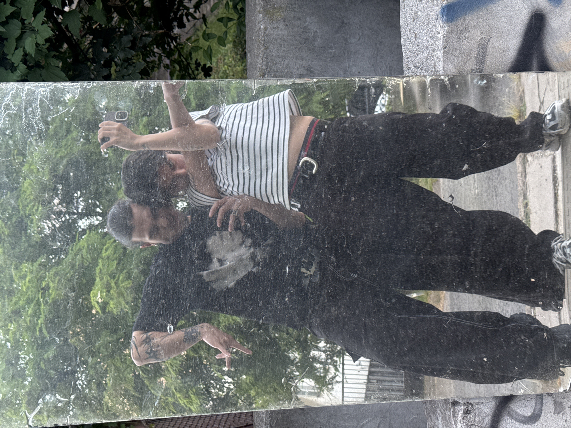 A full-body mirror selfie shows two individuals, one on the left and one on the right, reflected in a large, dirty mirror. The person on the left is wearing a black t-shirt with a graphic print and black pants. They have tattoos on their arm and are making a hand gesture. The person on the right is wearing a horizontally striped t-shirt with black pants and a belt. They are holding a phone to take the picture. Both individuals appear to be young adults. They are standing outdoors, with a blurred background of green trees and foliage, suggesting a park or natural area within a city. The ground beneath them appears to be paved. Graffiti is visible on a concrete structure to the right of the mirror. The overall impression is a casual, candid shot captured in an urban environment, possibly Sofia, Bulgaria, given the context provided.